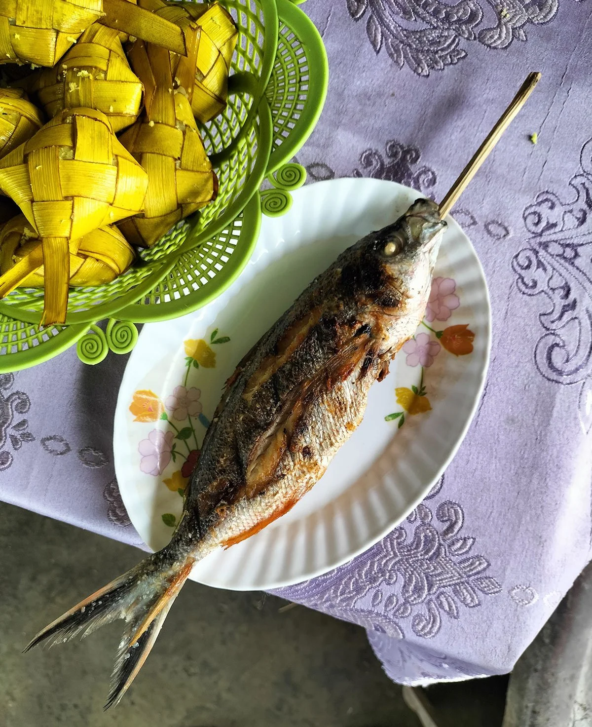Grilled fish on a floral patterned plate with a skewer, placed on a purple embroidered tablecloth.