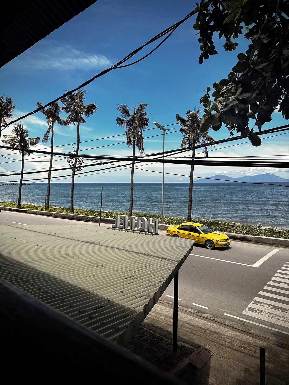 View of palm trees along the ocean, with a yellow taxi on the street, and a sign spelling 'HOTEL' next to the sidewalk, under a partly cloudy sky.