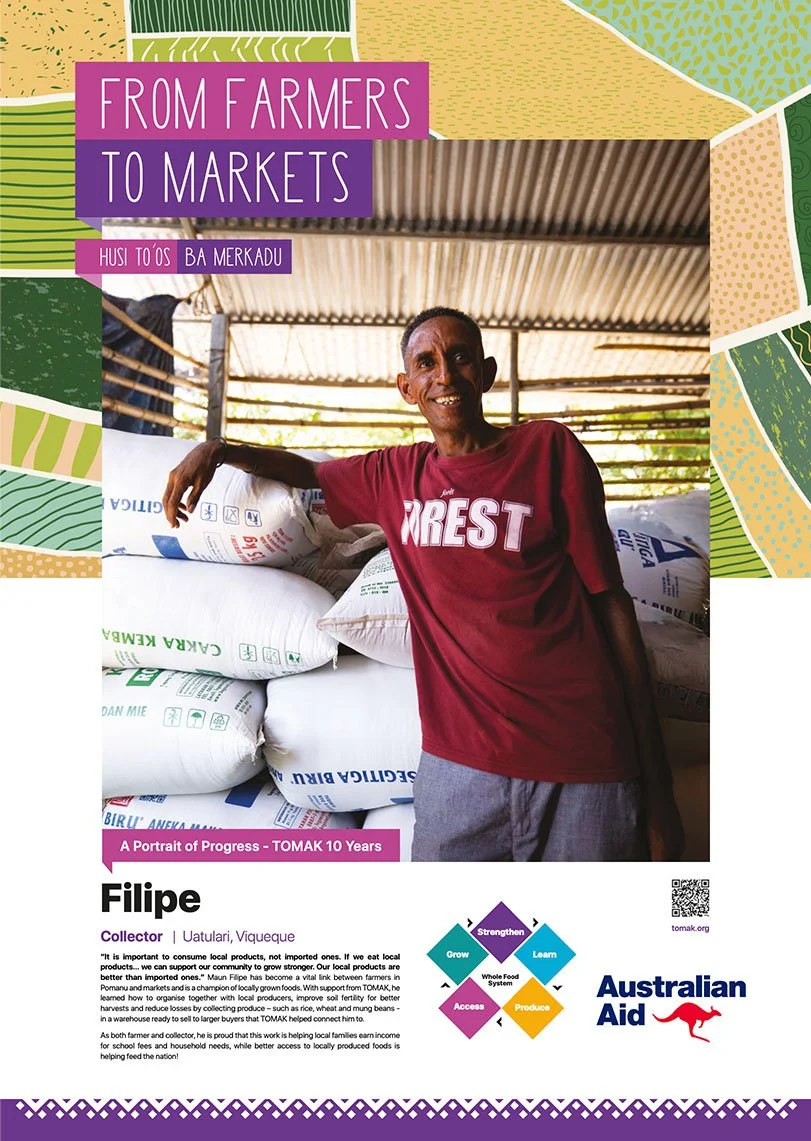 A smiling man standing in a warehouse with bags of local produce and a metal roof overhead. He wears a red t-shirt and gray pants.