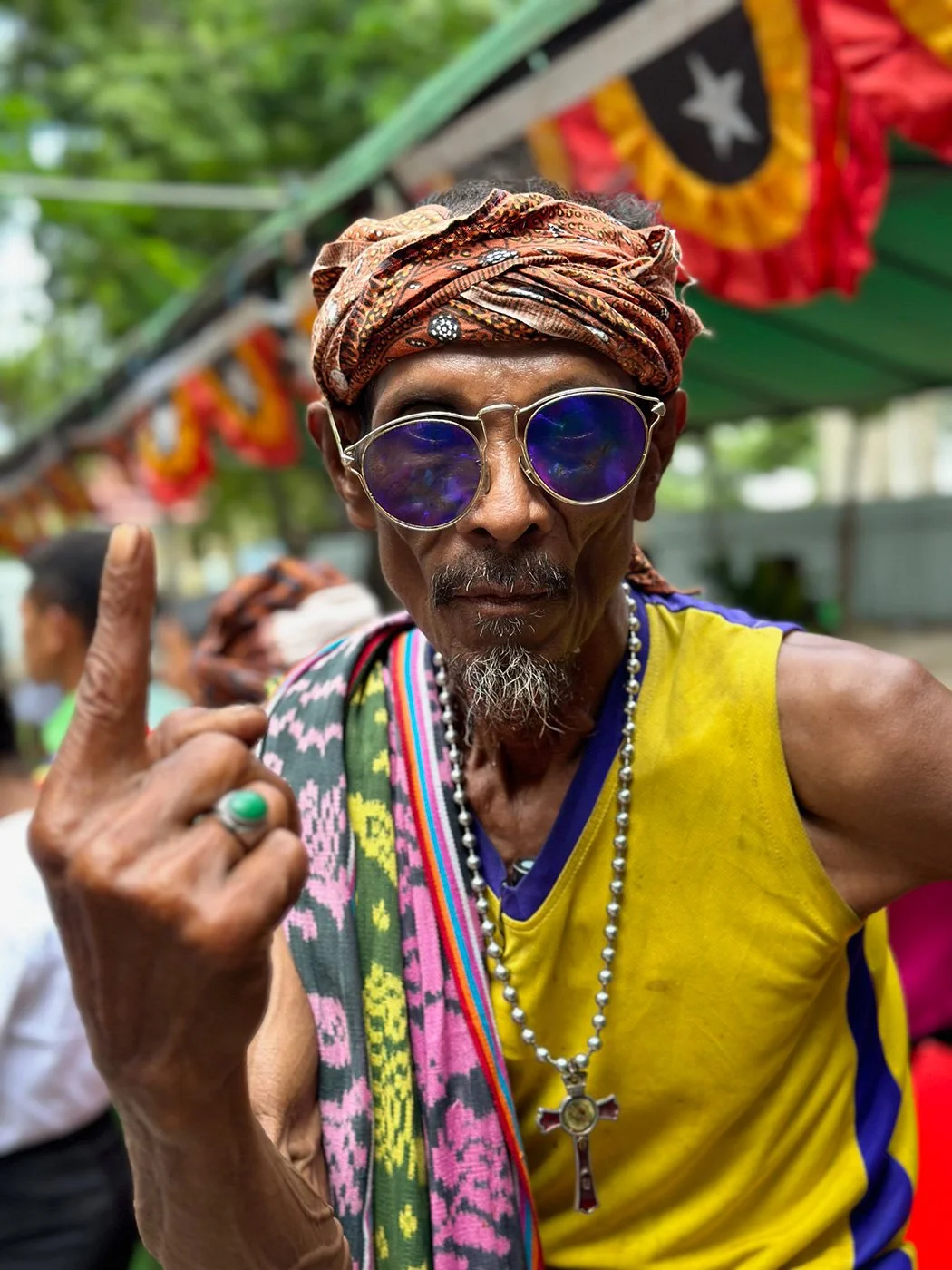An older man wearing sunglasses, a colorful headscarf, and yellow sportswear with purple trim. He has a beard, ring, and beaded necklace with a cross pendant. He is raising his left index finger, and there are colorful banners in the background, sugg