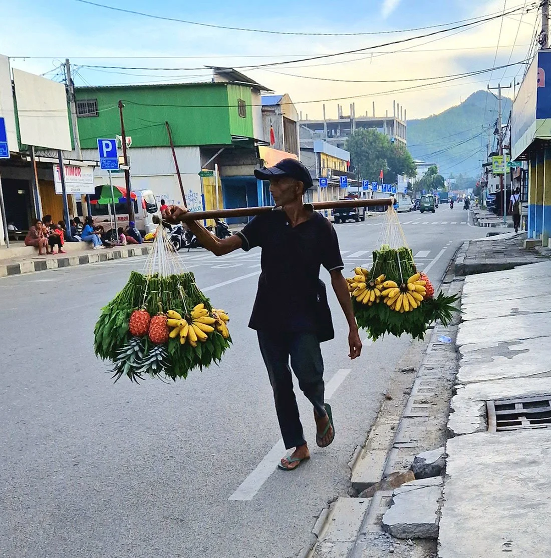 A street vendor walks on the road carrying two baskets of fruit, including bananas, pineapples, and leafy greens, balanced on a pole across his shoulder, with a city street and people in the background.