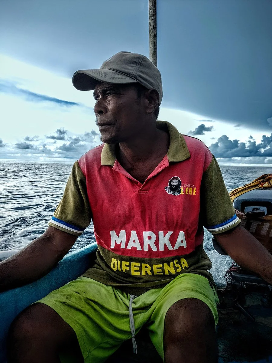 A man wearing a khaki cap and a colorful polo shirt with logos and text, sitting on a boat with the ocean and cloudy sky in the background.