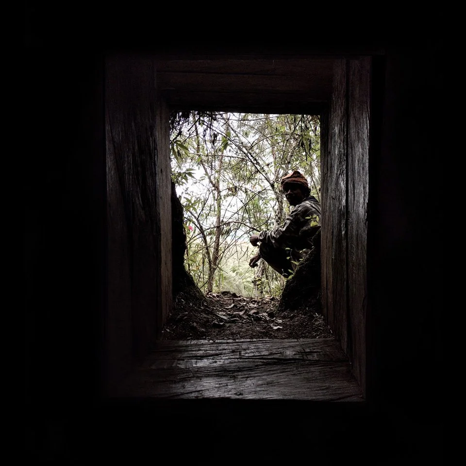 Person squatting outdoors near trees and bushes, viewed through a square opening from inside a dark structure.