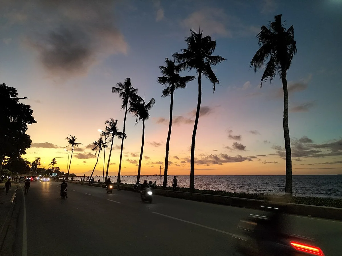 Sunset over a beach with tall palm trees, a road with vehicles, and people walking along the shoreline.