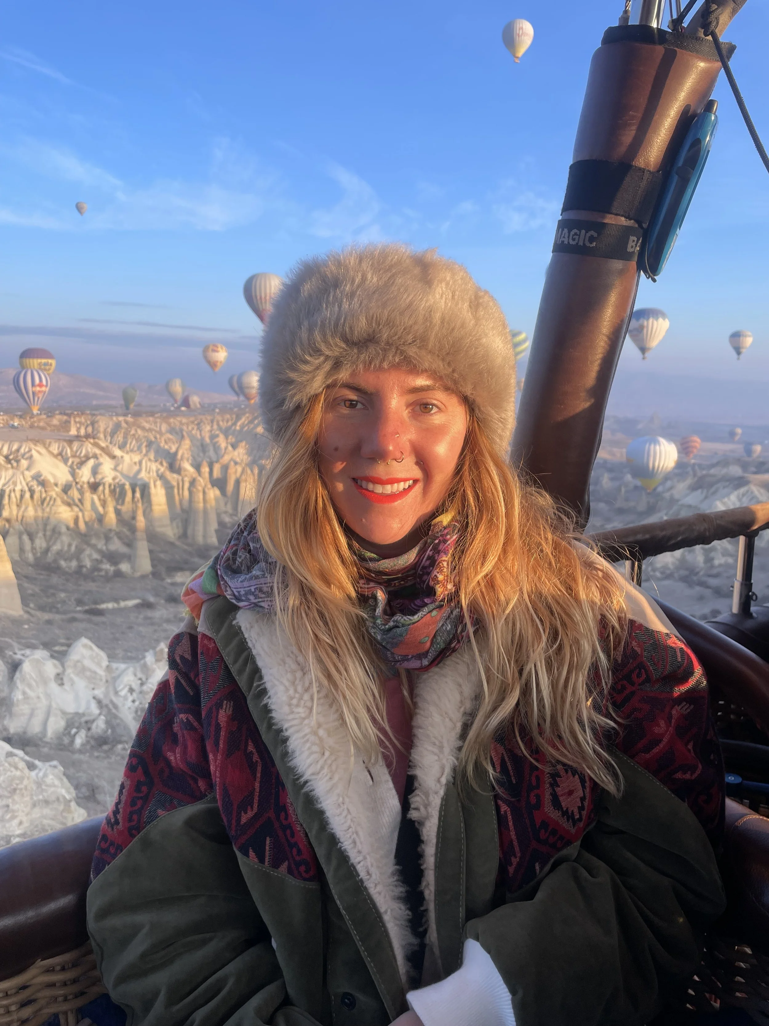 A woman with long hair, wearing a fluffy hat and a colorful patterned jacket, smiling in a hot air balloon with the landscape of Cappadocia and several other hot air balloons in the sky in the background.