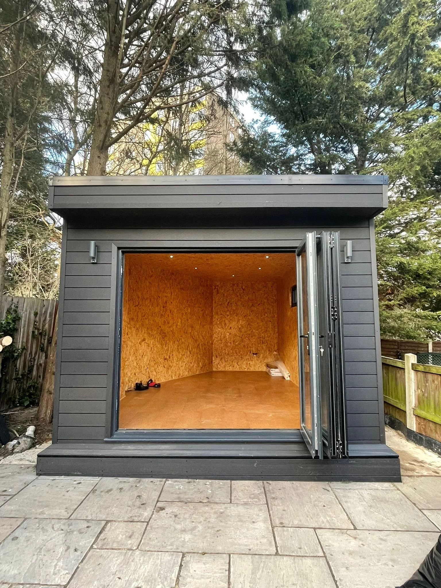 A small gray shed with an open door, revealing an unfinished wooden interior, situated on a stone patio surrounded by trees and a wooden fence.