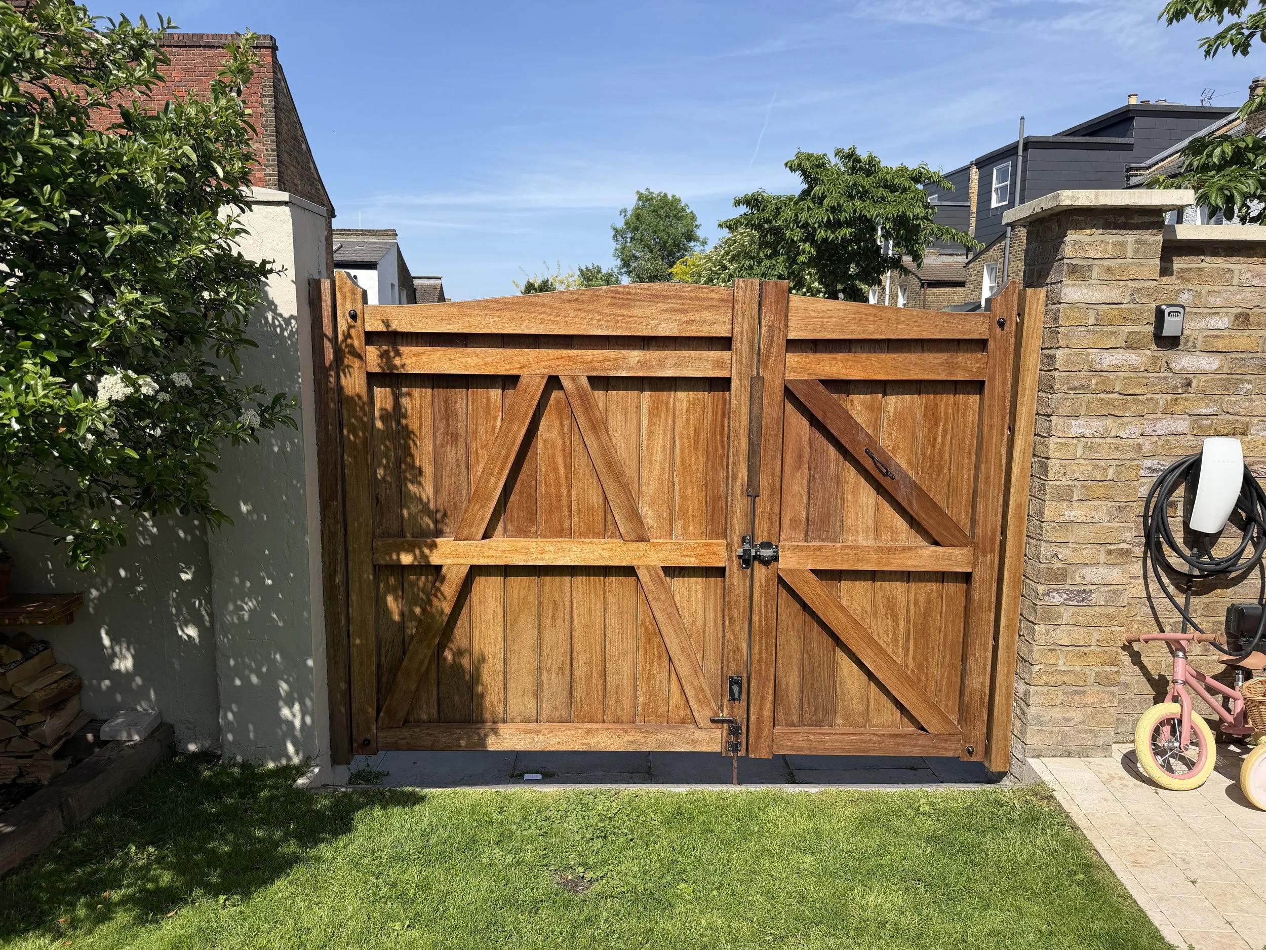 A wooden garden gate with black hinges and latch in front of a brick and white wall, with trees and rooftops in the background under a blue sky.