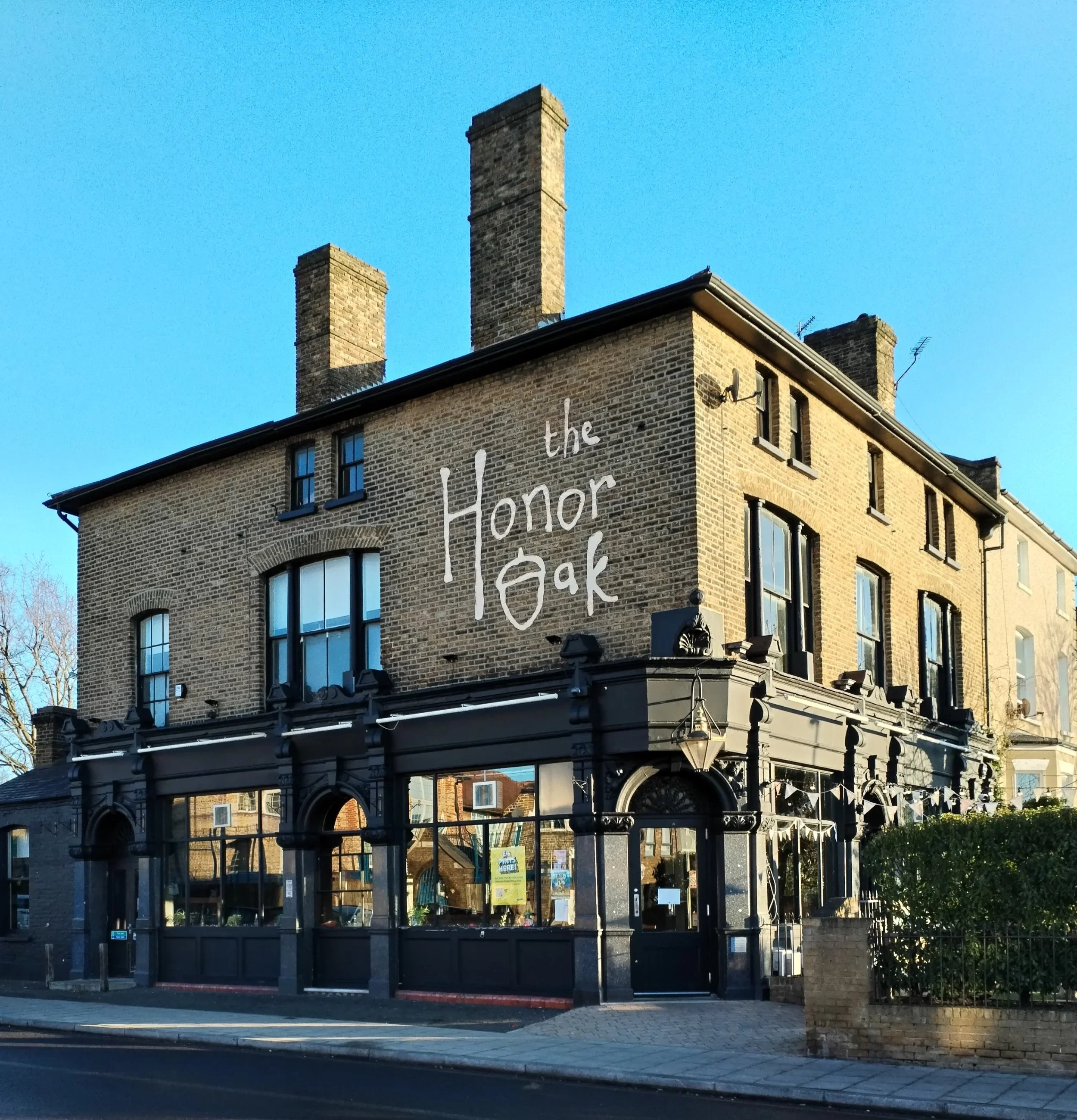 A brick building with large windows and black lanterns on the exterior, displaying the words 'Honor the' painted on the upper part of the brick wall. The building has several tall brick chimneys and some antennas on the roof, set against a clear blue sky. The lower part includes a black windowed structure resembling a bus stop or storefront with an assortment of posters and signs visible through the windows.