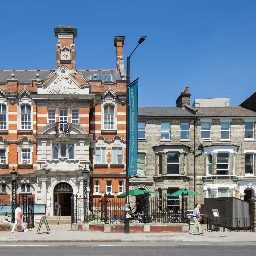Building facades along a city street with a South London Gallery banner, outdoor seating with green umbrellas, people walking, and a clear blue sky.