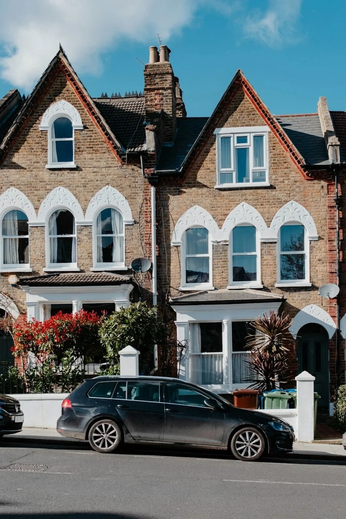 A red brick townhouse with decorative white trim around the windows, a small garden with shrubs and trees, and two cars parked on the street in front.