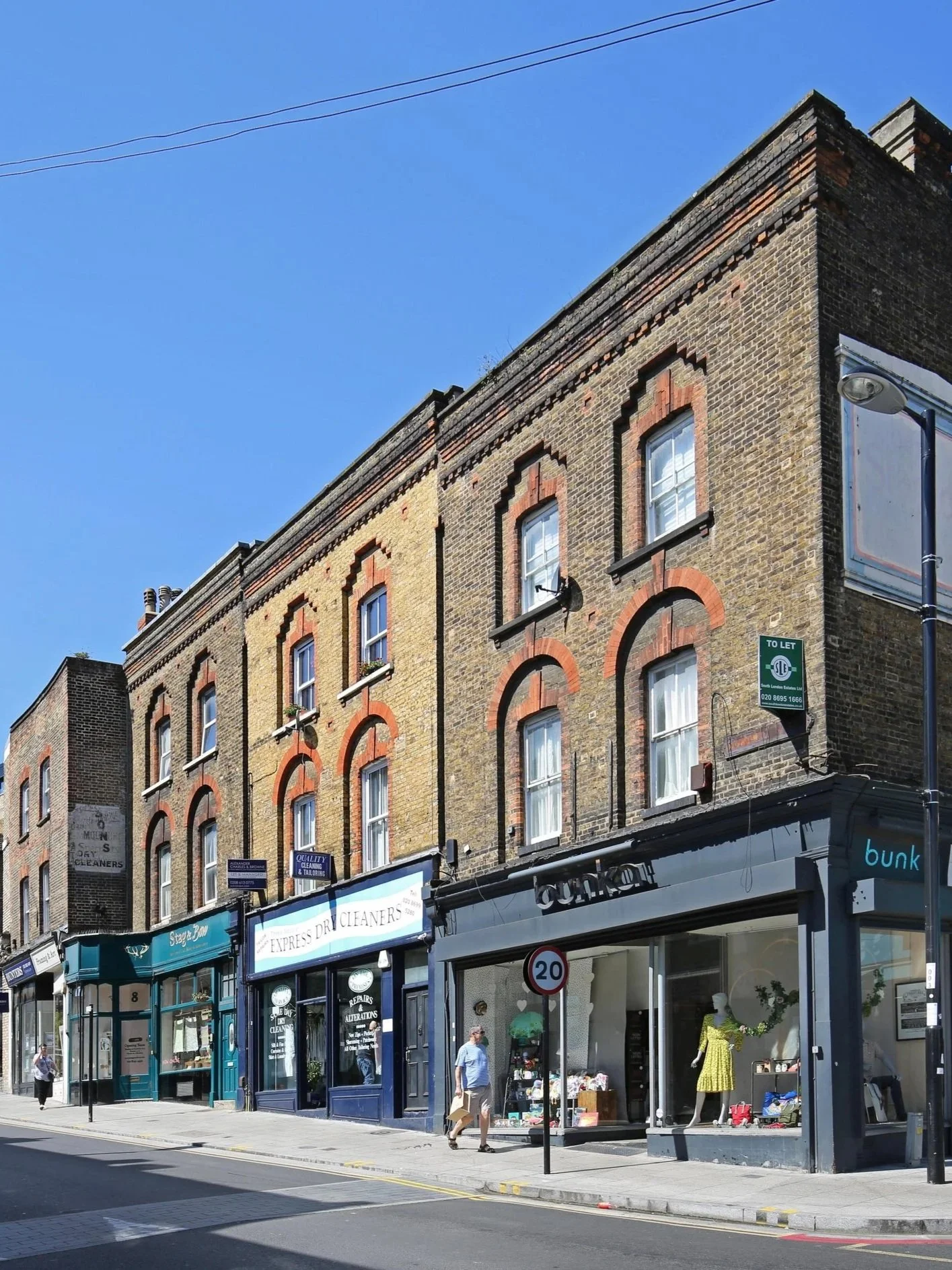 Street view of a row of old brick buildings with retail shops on the ground floor, including a dry cleaner, and people walking on the sidewalk under a clear blue sky.
