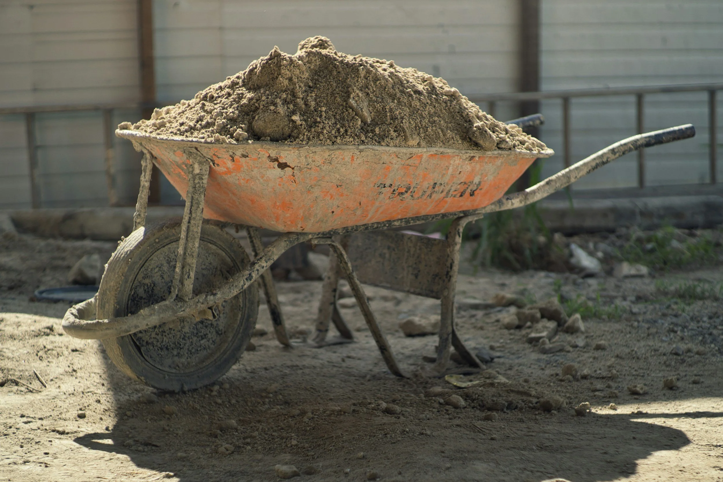 An orange wheelbarrow filled with gray soil or sand, positioned on uneven ground outdoors near a metal fence.
