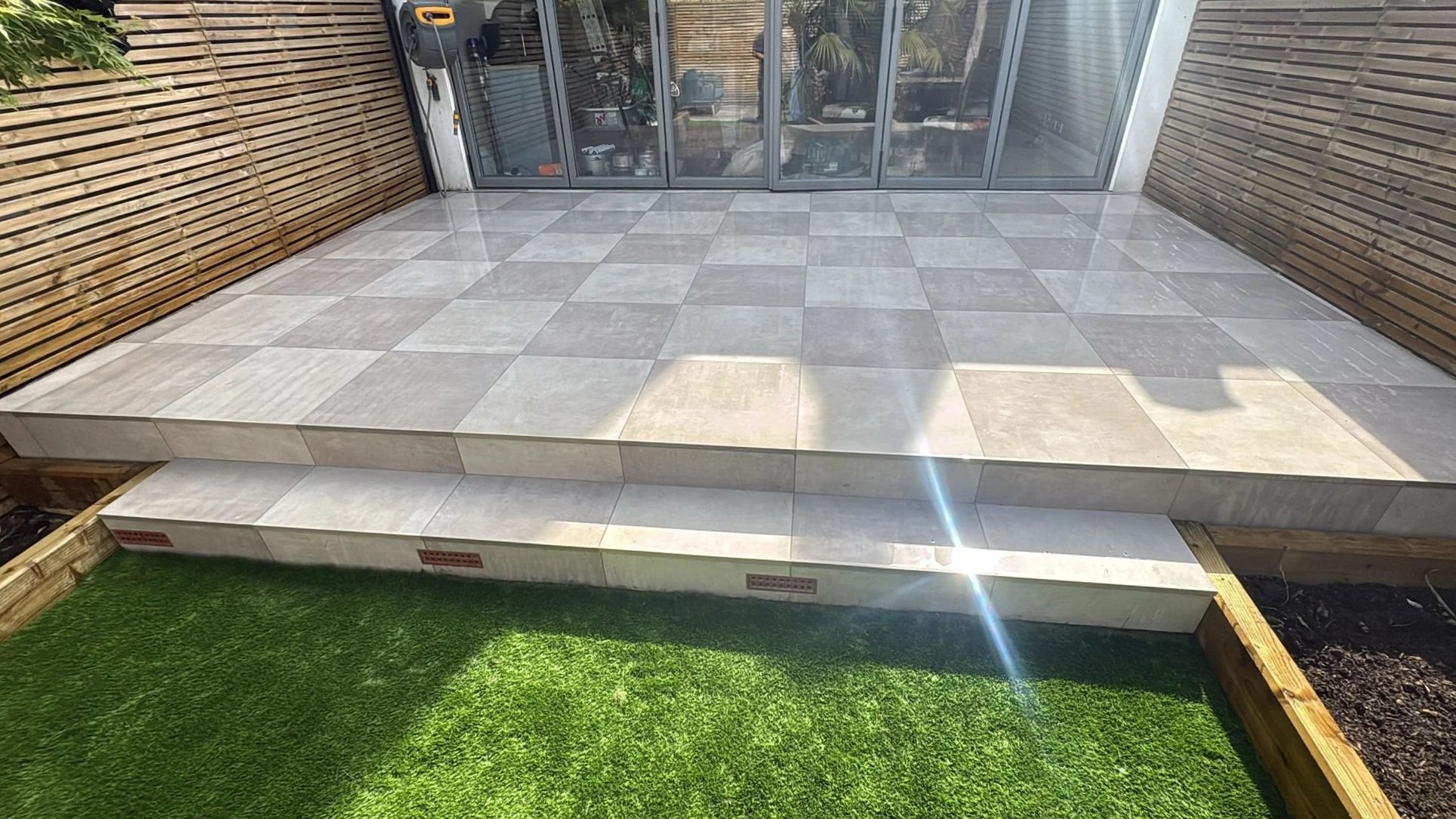 A newly built tiled patio with steps leading up to sliding glass doors, surrounded by a wooden fence on two sides and artificial grass on the ground.