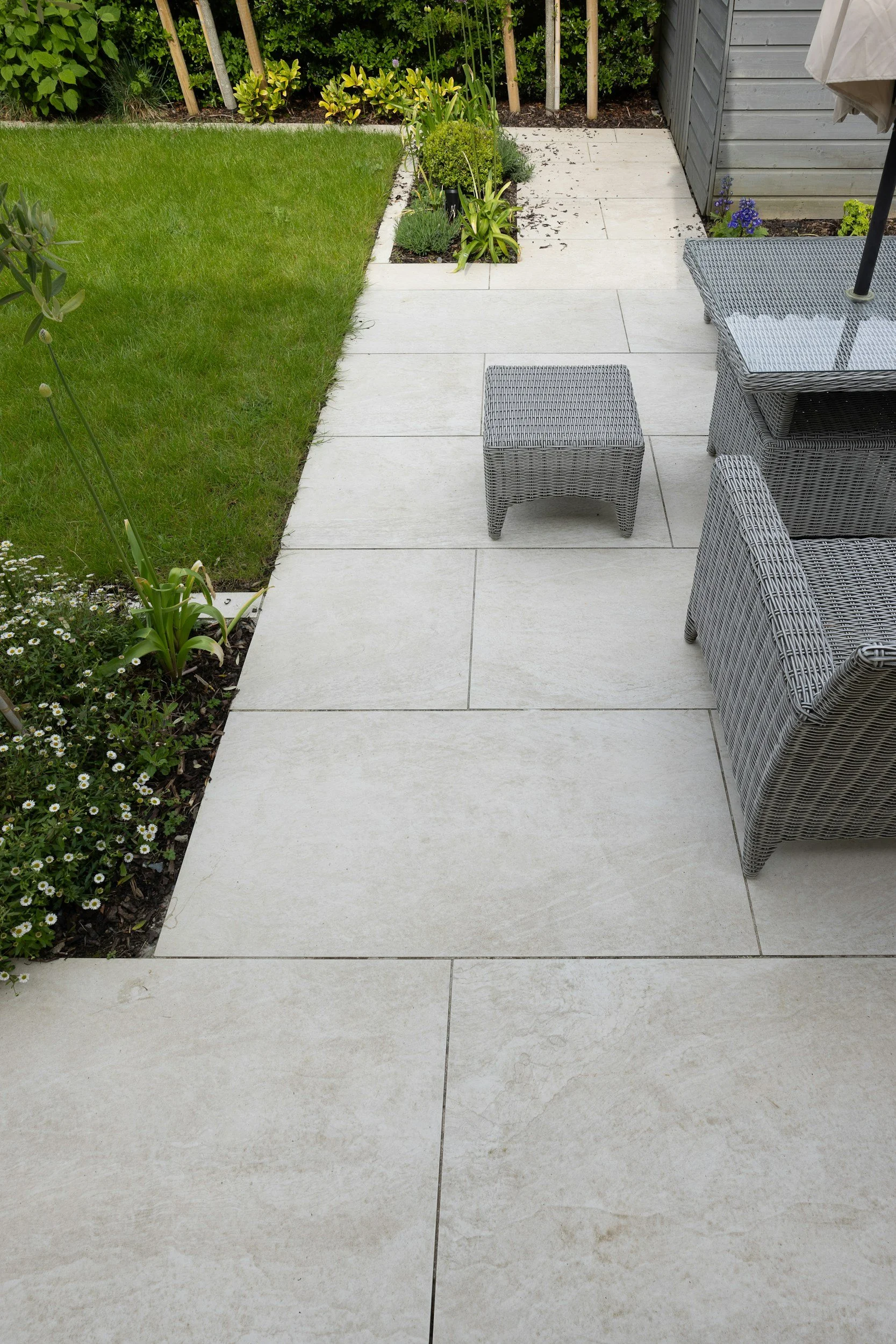 A backyard patio area with large light-colored tiles, outdoor furniture including a wicker chair and small matching table, a lawn on the left, a garden bed with plants, and a gray wooden shed on the right.