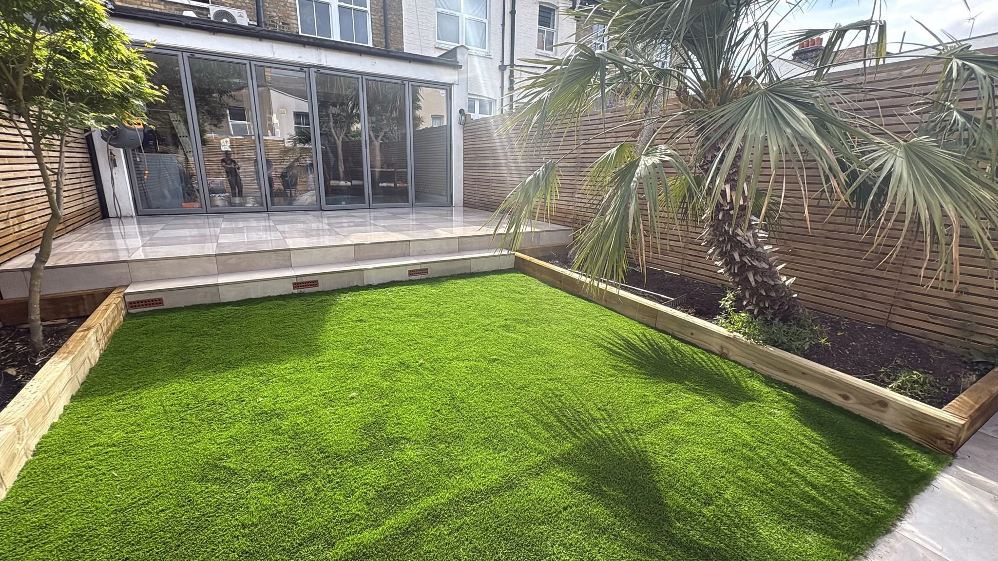 A backyard with a green artificial grass lawn, a raised stone patio area, a wooden fence, a large palm tree, and a glass extension on the house.