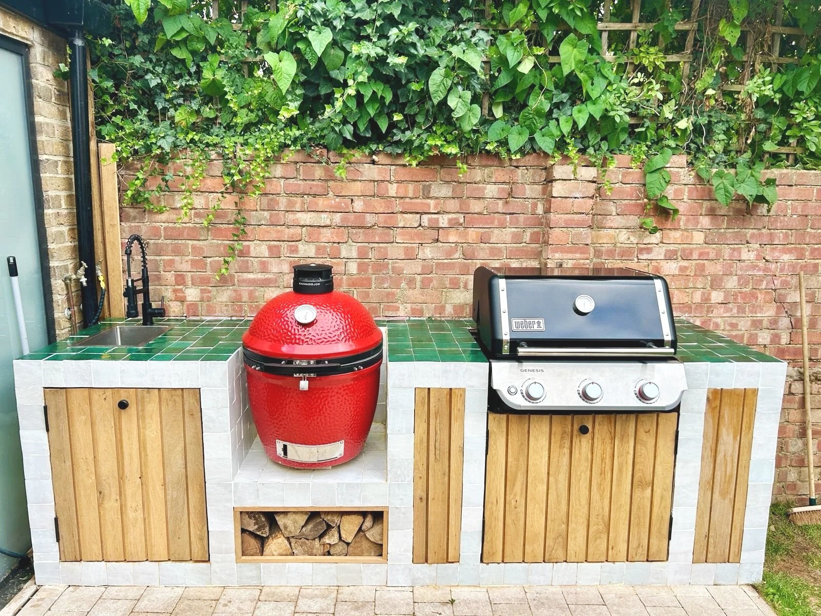 Outdoor kitchen with a small sink, a red smoker grill, and a black gas grill, set against a brick wall and greenery.