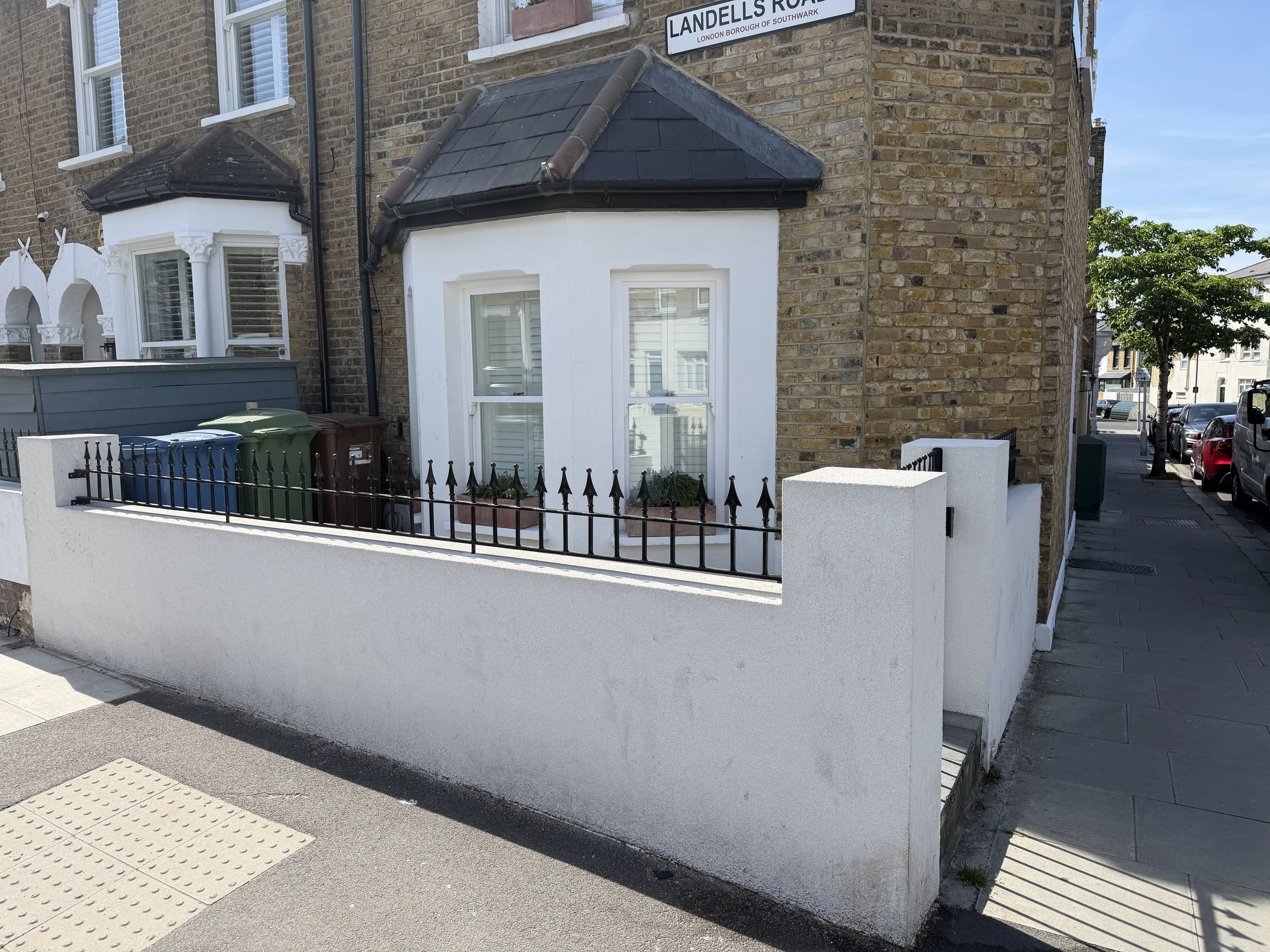 A street corner with a brick building labeled 'Landells Road' and a white wall with black wrought iron fence, trash bins, and a sidewalk, under a clear blue sky.