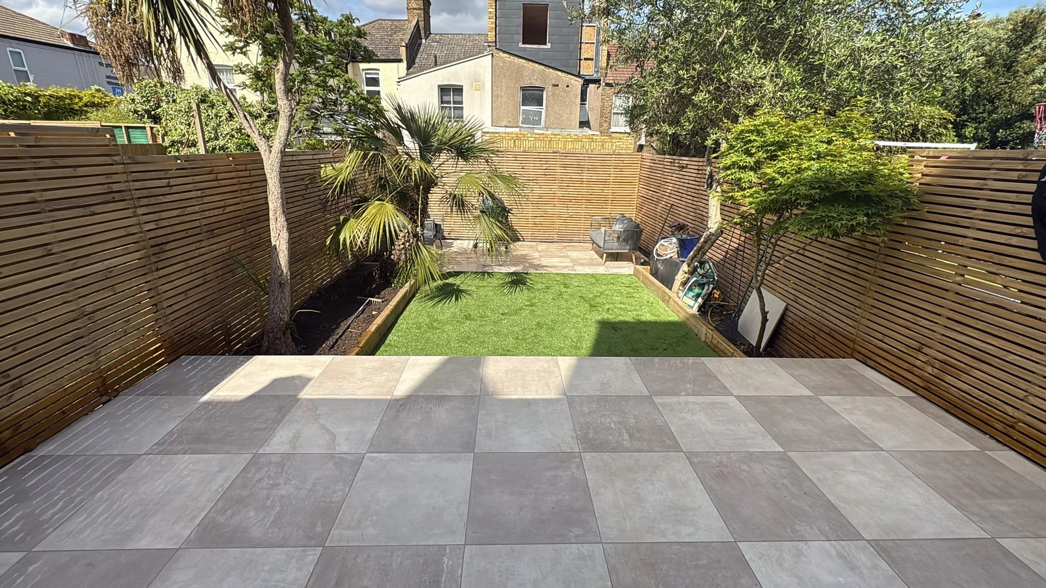 A backyard with a tiled patio, wooden privacy fence, small grassy area, and various trees and plants, with neighboring buildings visible in the background.