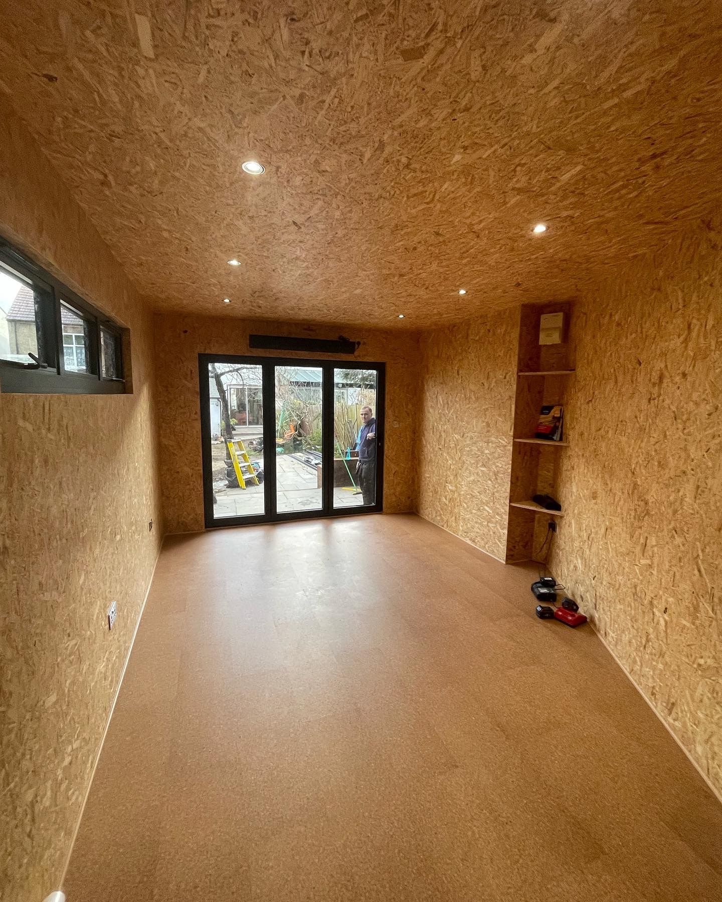 Empty interior room with OSB panel walls and ceiling, a sliding glass door at the back, a small shelf on the right wall, and some tools on the floor.
