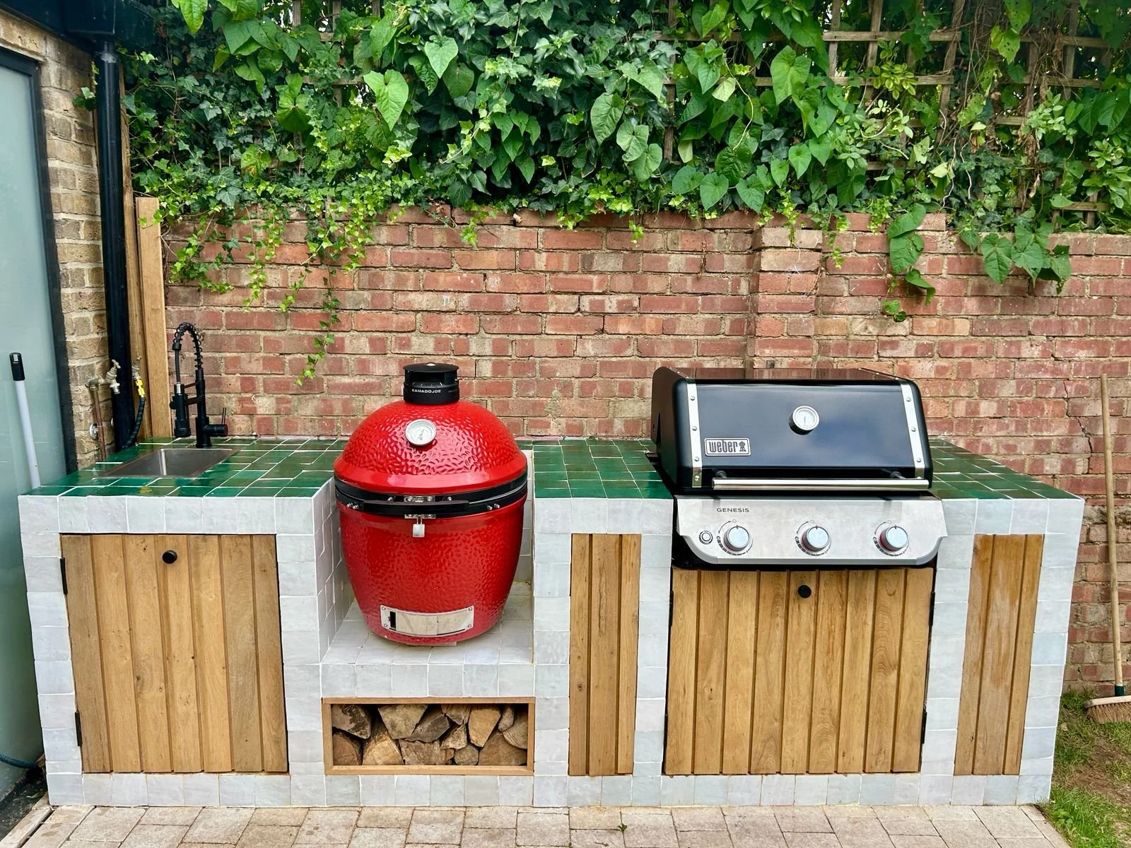 An outdoor kitchen setup with a green tiled countertop, a red Kamado-style grill, a black Weber gas grill, a small sink with a black faucet, and storage space beneath the counter filled with firewood, all against a brick wall with green foliage overh
