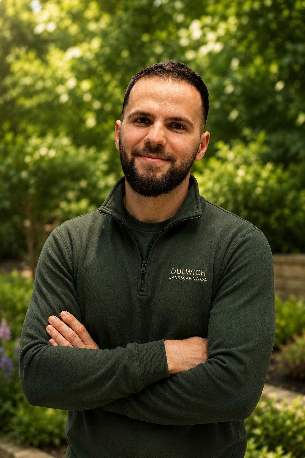 A man with a beard, smiling, with arms crossed, wearing a dark green jacket with 'DULWICH LANDSCAPING CO.' logo, standing outdoors with lush green trees and plants in the background.