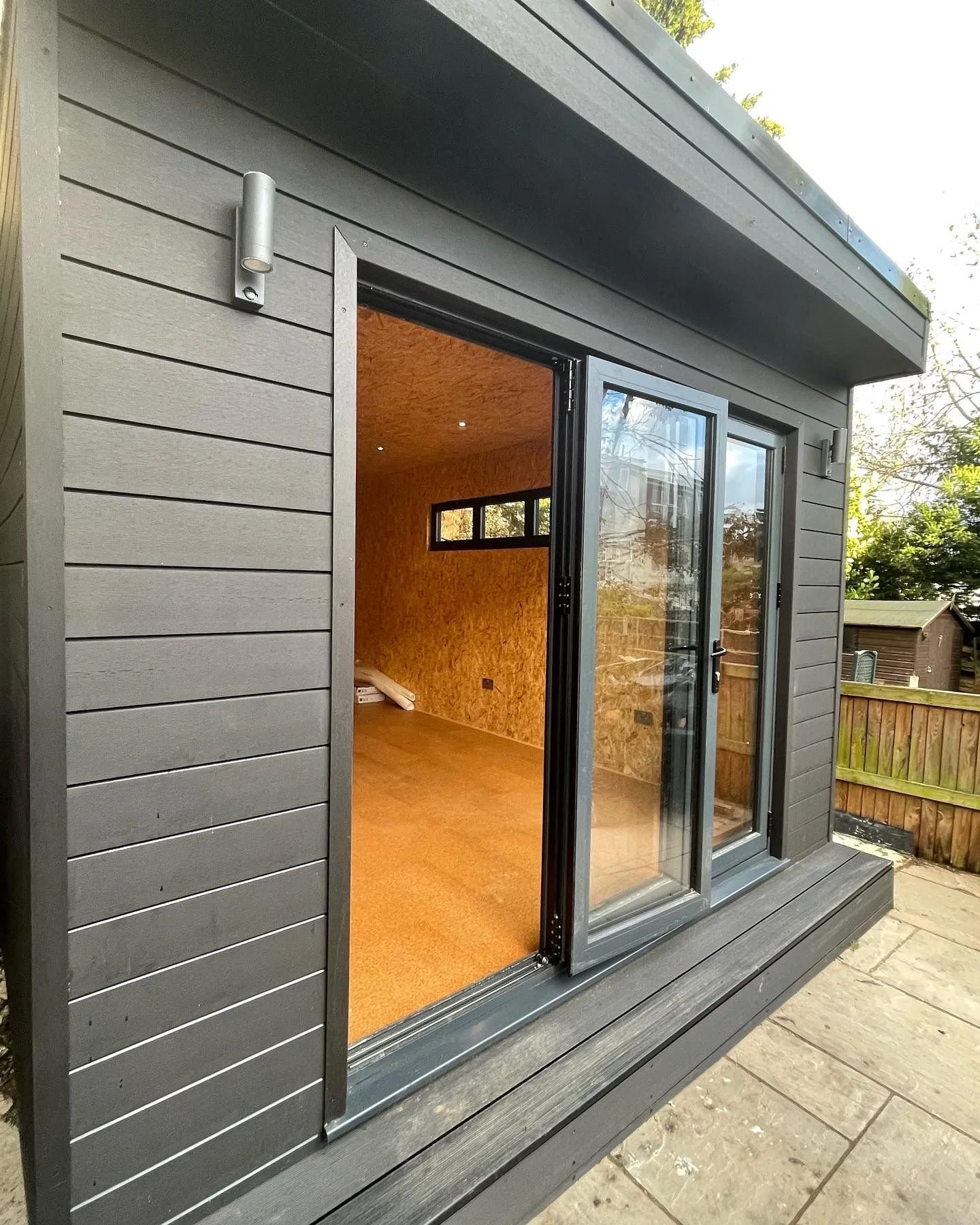 A modern exterior door opens to reveal a cozy interior with wood-paneled walls and a small window, set against a gray siding exterior with outdoor lighting fixtures and a wooden deck.