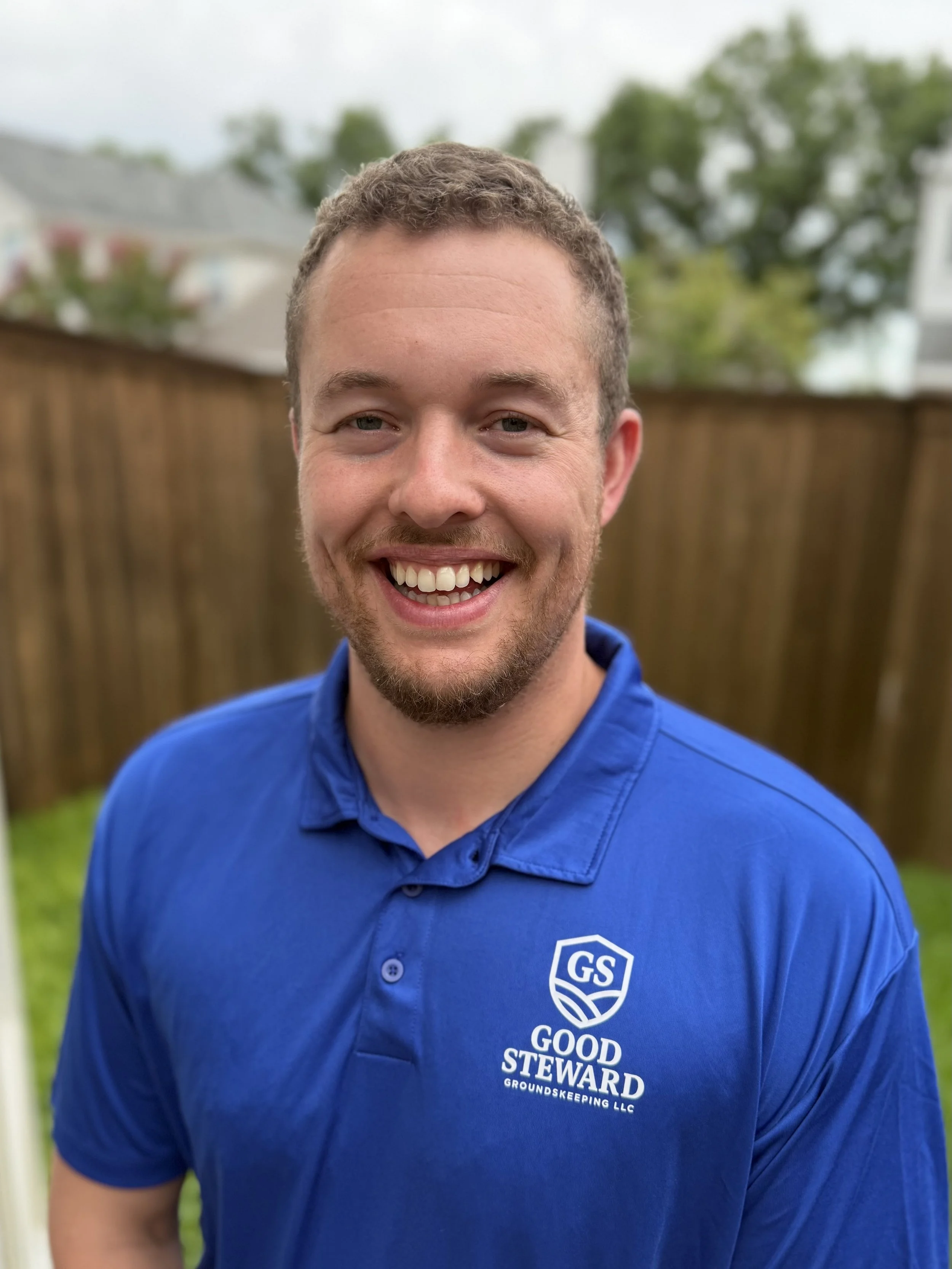 A smiling man with short curly hair and facial hair, wearing a blue collared shirt with a logo that says 'GS Good Steward Groundskeeping LLC', standing outside in front of a wooden fence and green trees.