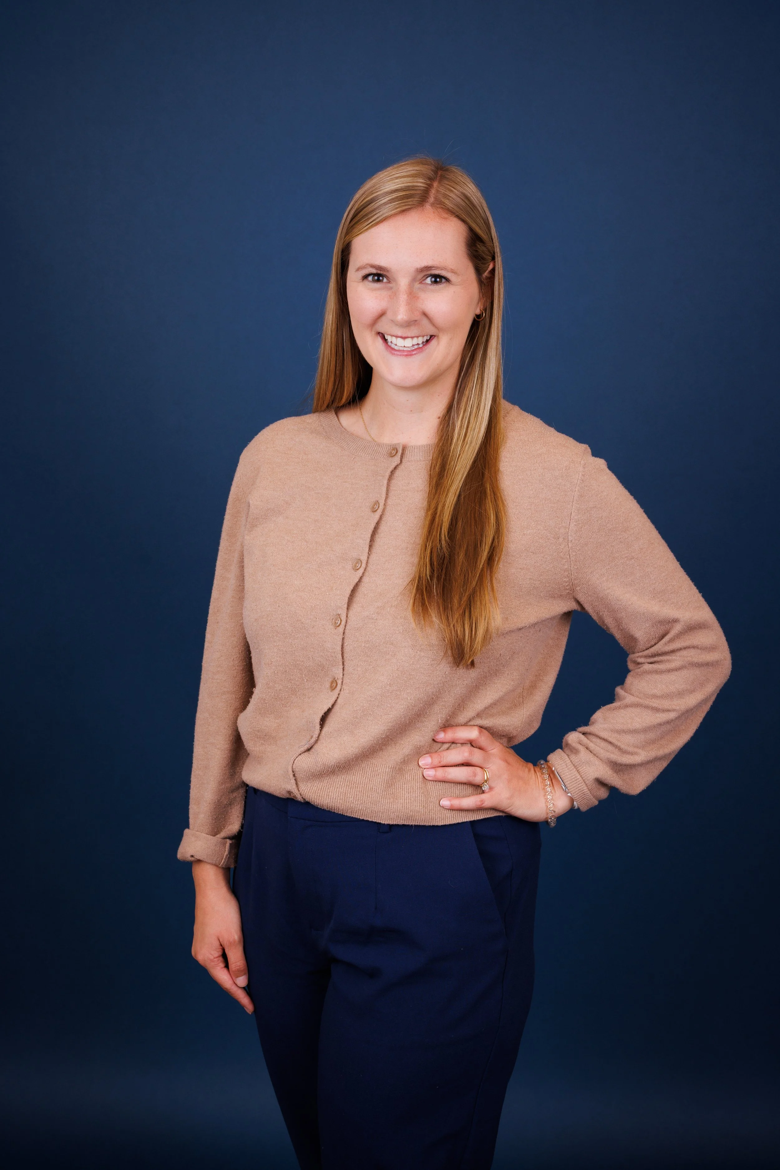 Portrait of a woman with long light brown hair, wearing a beige button-up sweater and navy pants, posing with one hand on her hip against a blue background, smiling.