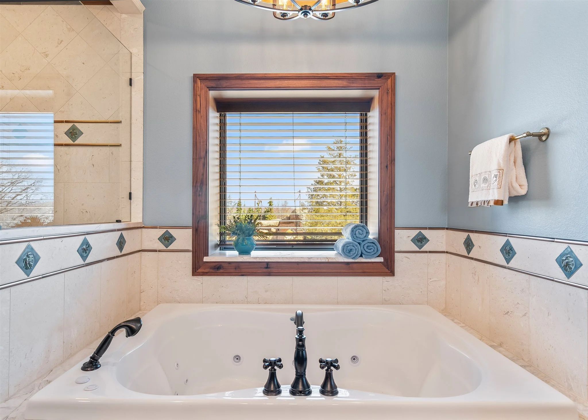 A bathroom with a large soaking bathtub, a window with blinds, a towel rack with a towel, and a decorative vase with flowers by the window, viewed from the bathtub side.