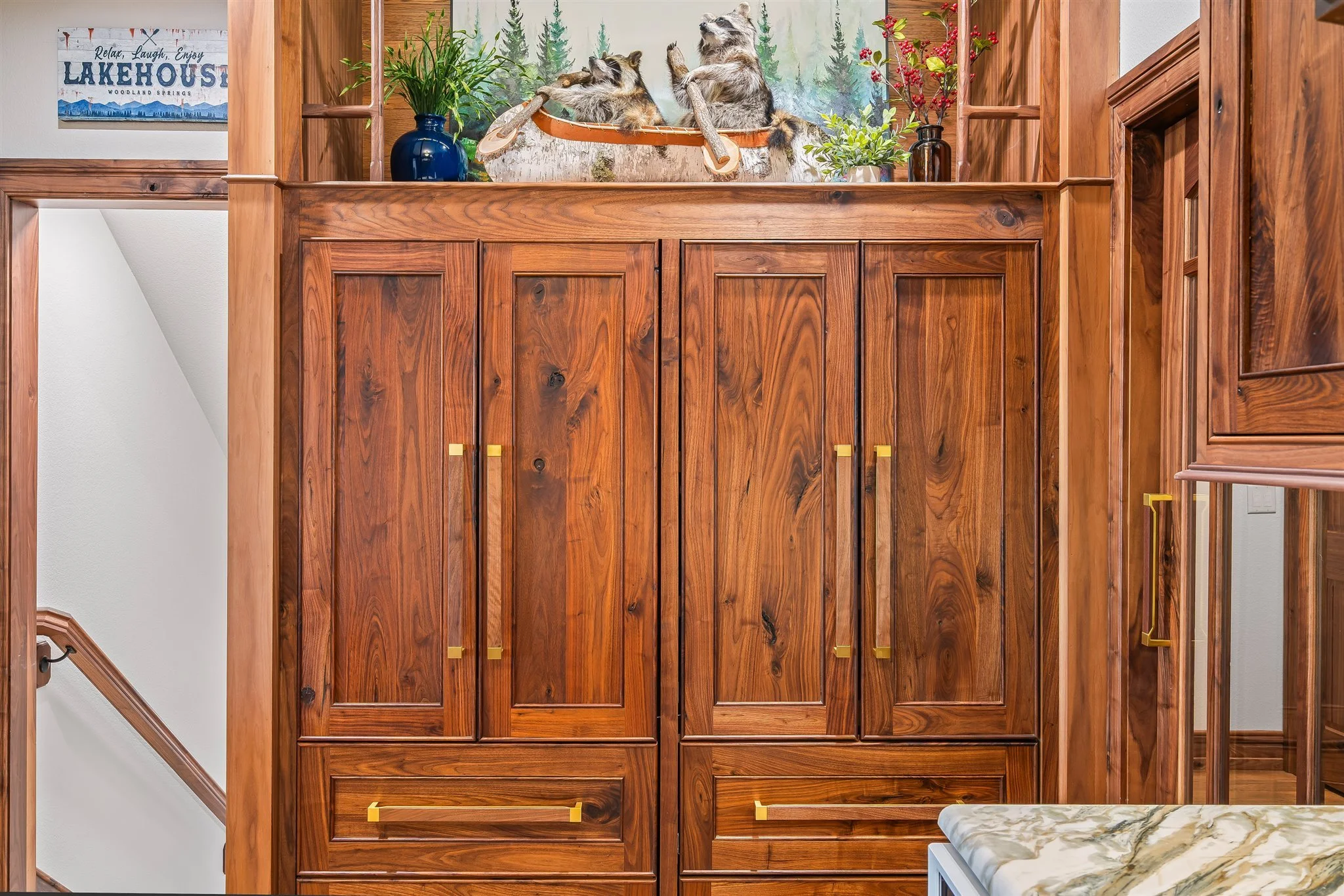 large wooden cabinet with gold handles in a room with white walls, decorated with potted plants and a framed sign reading 'Relax, Laugh, Enjoy LAKEHOUSE Woodlands Springs', above a staircase with a wooden railing.