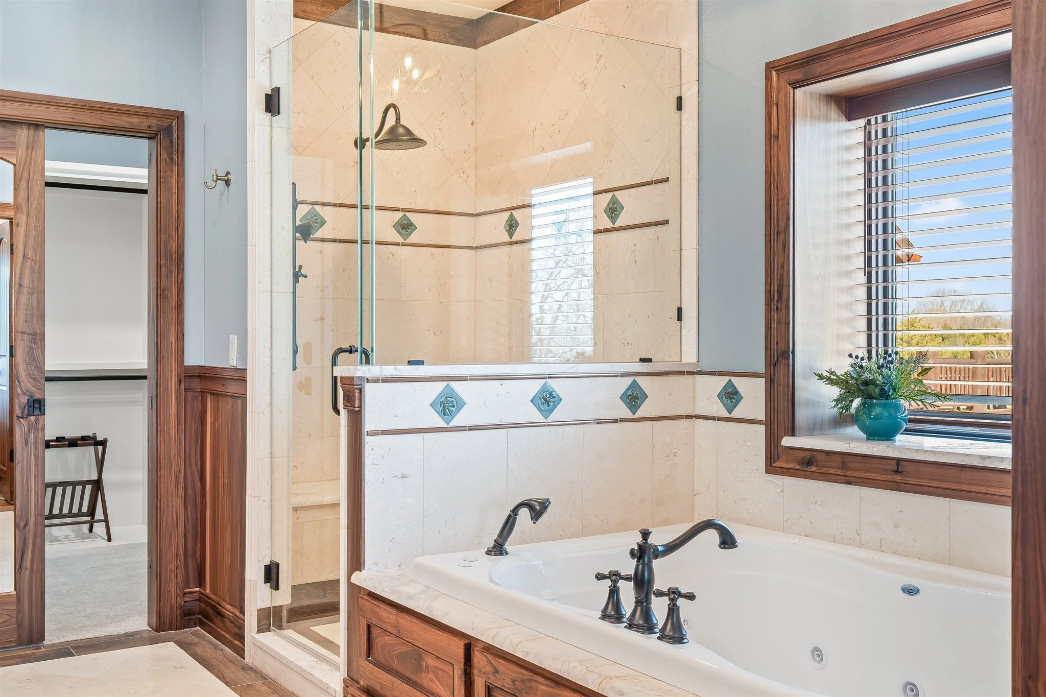 Bathroom with a built-in bathtub, a double sink vanity with black fixtures, a walk-in shower with glass doors, wood framed window with blinds, and a shelf with a teal vase holding flowers.
