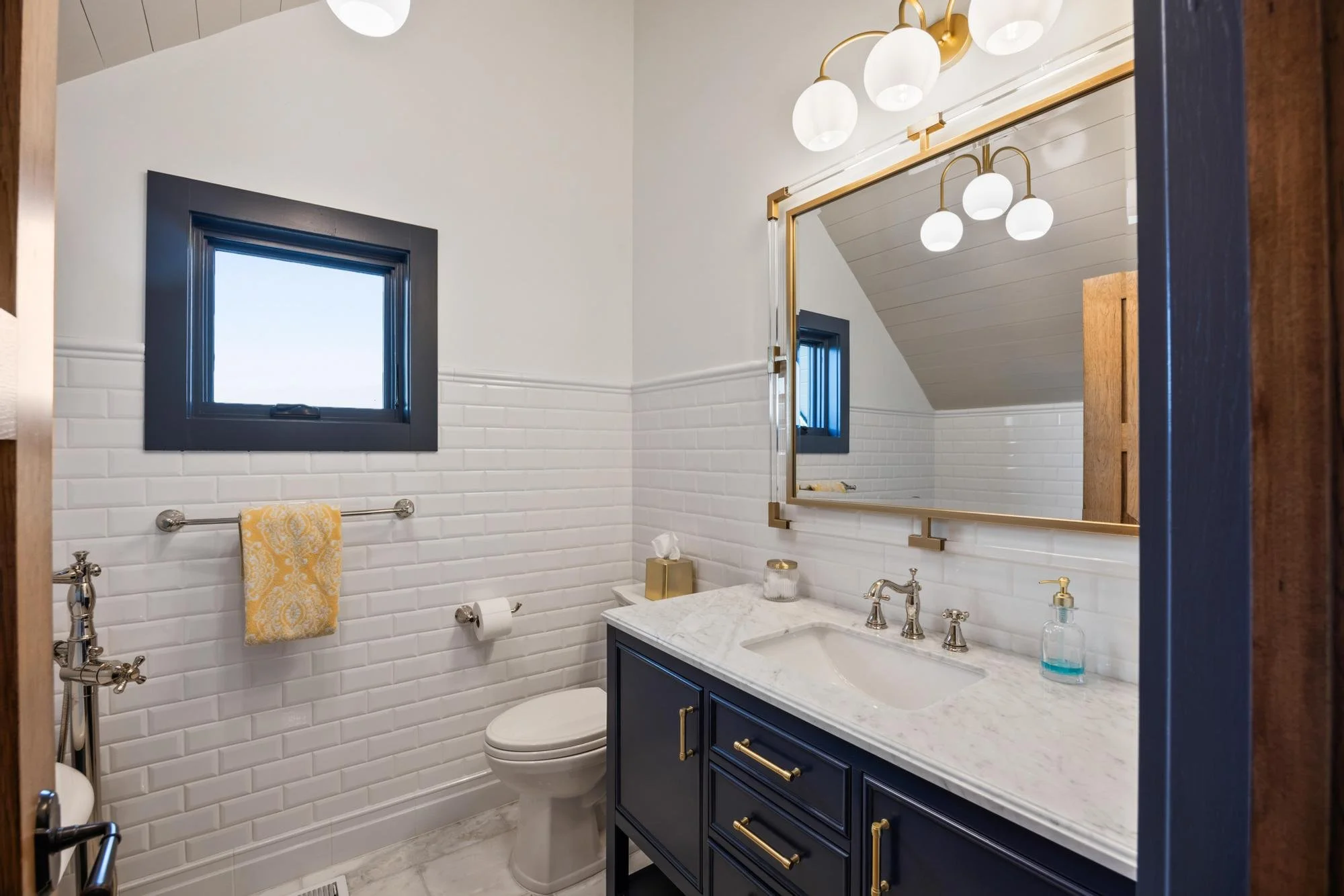 Bathroom with white subway tile walls, a navy blue vanity with gold accents, a marble countertop, a white sink, and a large mirror with gold trim. There are two small windows with dark frames, a gold tissue box, and a yellow patterned towel hanging o