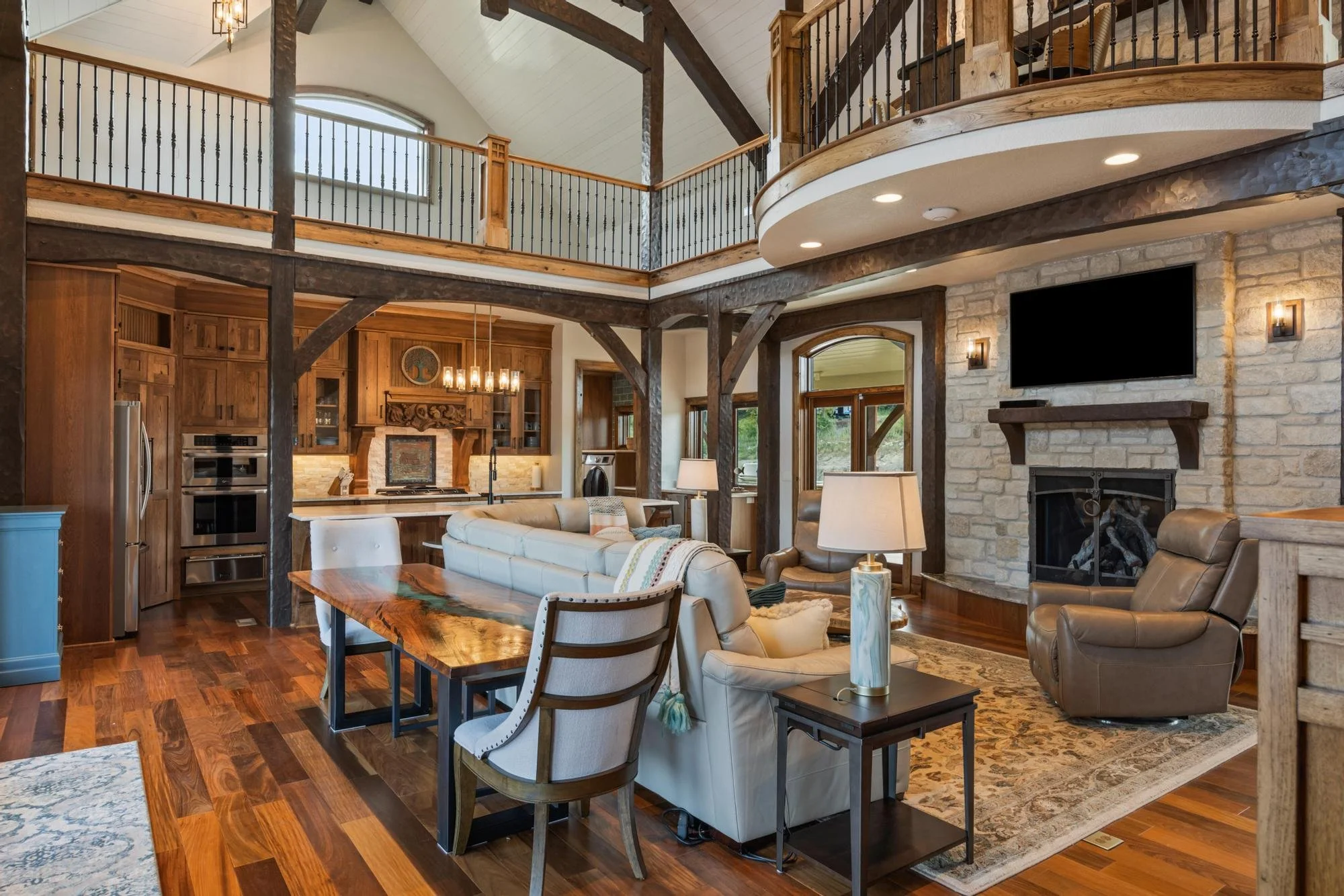 Living room with wood and stone accents, a flat-screen TV above the fireplace, beige sofas, a wooden coffee table, and large windows.