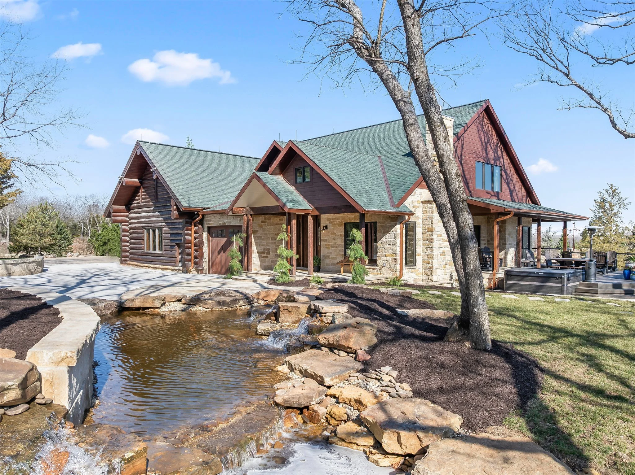 A house with wooden and stone exterior, green roof, and large windows, situated beside a small pond with rocks, surrounded by trees and a clear blue sky.