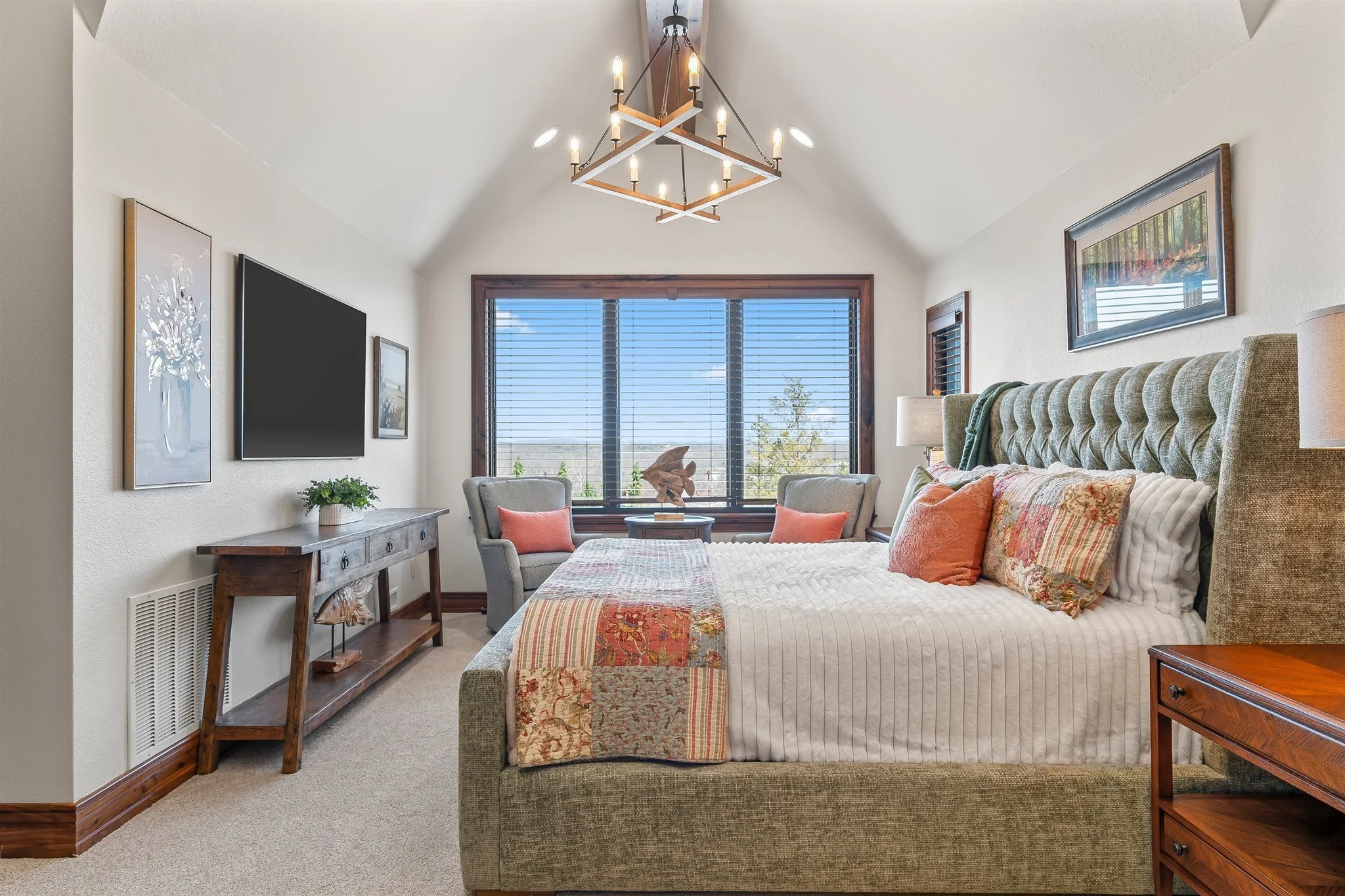 A cozy bedroom with a large bed with a textured headboard, patterned bedspread, and decorative pillows. There's a sitting area with two armchairs and a small table, large window with blinds, a dresser with a potted plant, and wall-mounted paintings. 