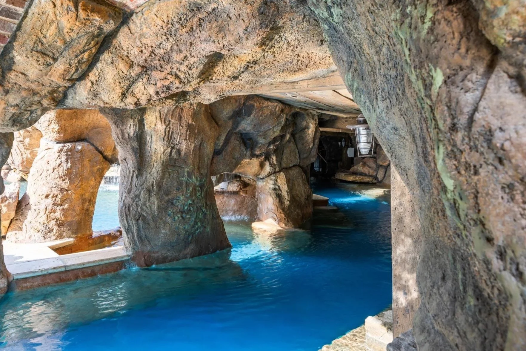 Indoor swimming pool area with artificial rock formations and a small waterfall, viewed through a rocky archway.