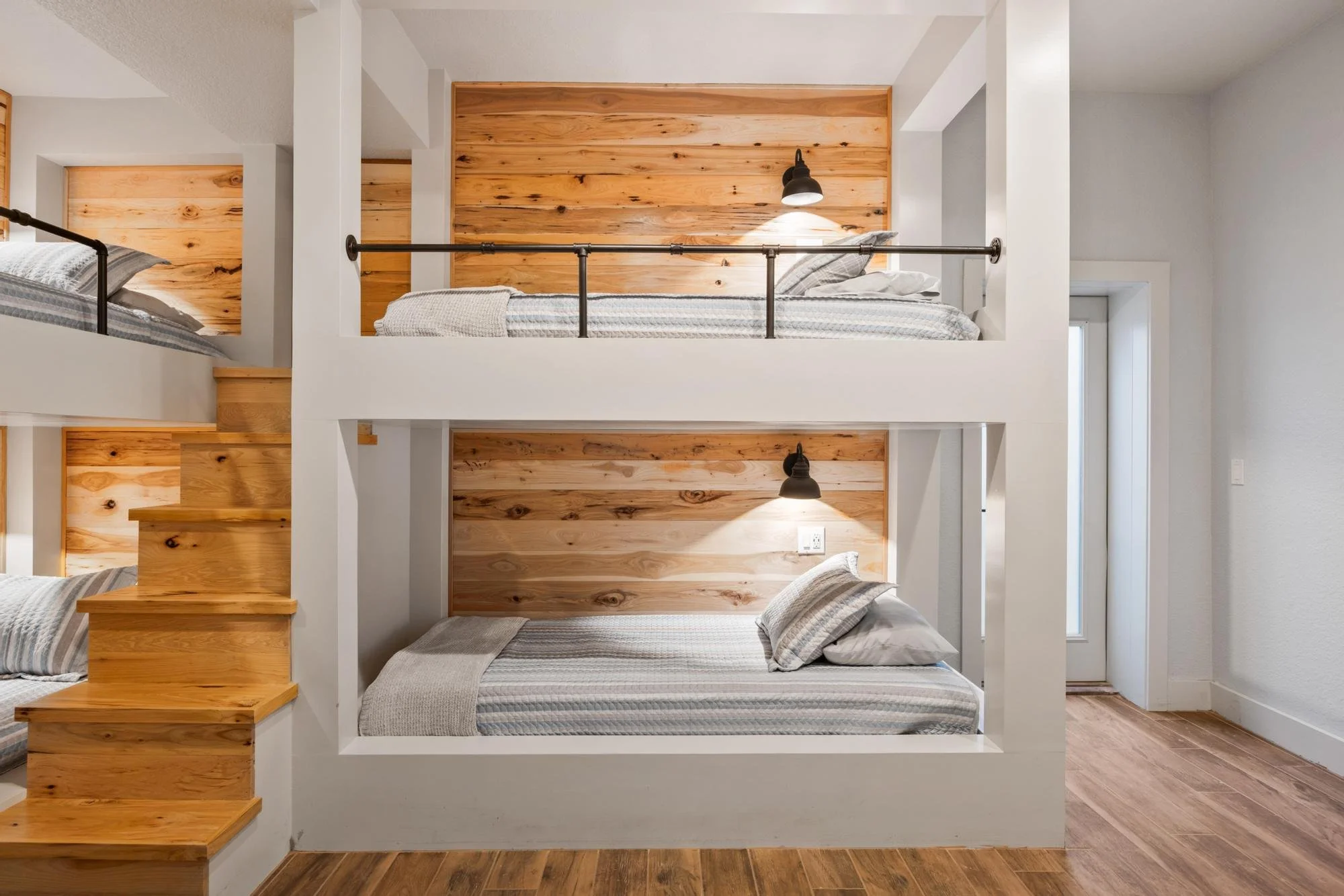 Multiple beds in a modern bedroom with wood accents, including a partial staircase, white walls, and black wall-mounted lamps.