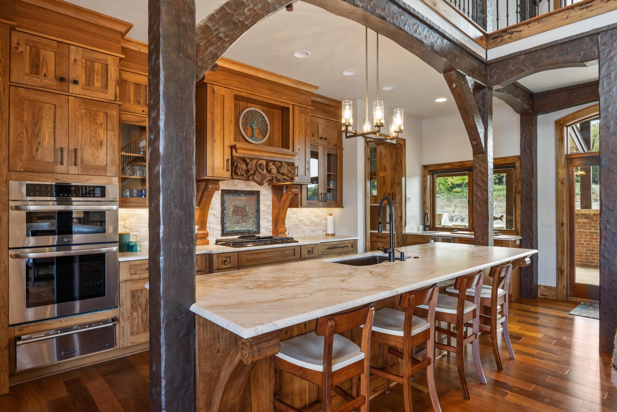 Kitchen with wooden cabinets, marble island, hanging light fixture, large windows, and a stone fireplace with wood details.