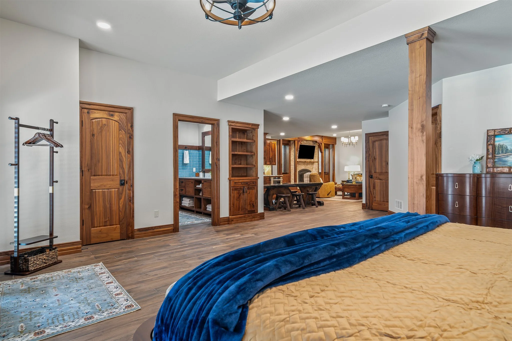Interior view of a bedroom with a partially visible bed covered in a yellow quilt and a blue blanket. The room has wooden doors, trim, and shelves, with a living space in the background featuring a fireplace, television, and seating area. The floorin