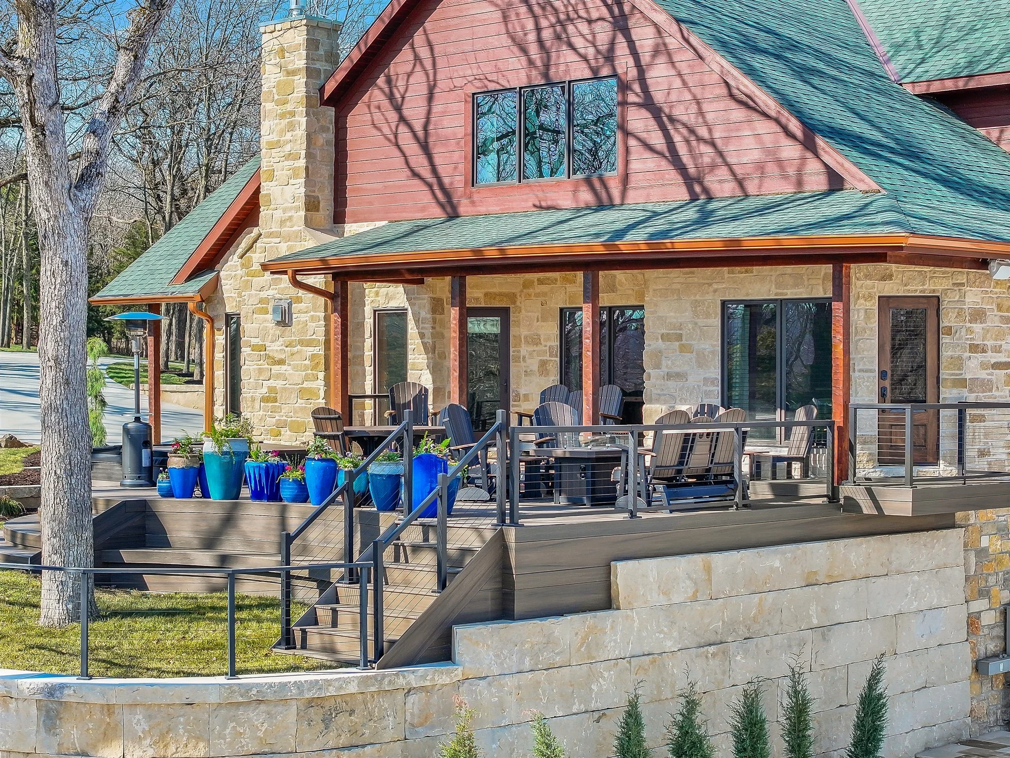 A two-story house with a stone and red wood exterior, green shingle roof, large windows, and an outdoor patio with furniture and potted plants. There are trees casting shadows, and the sky is clear and blue.