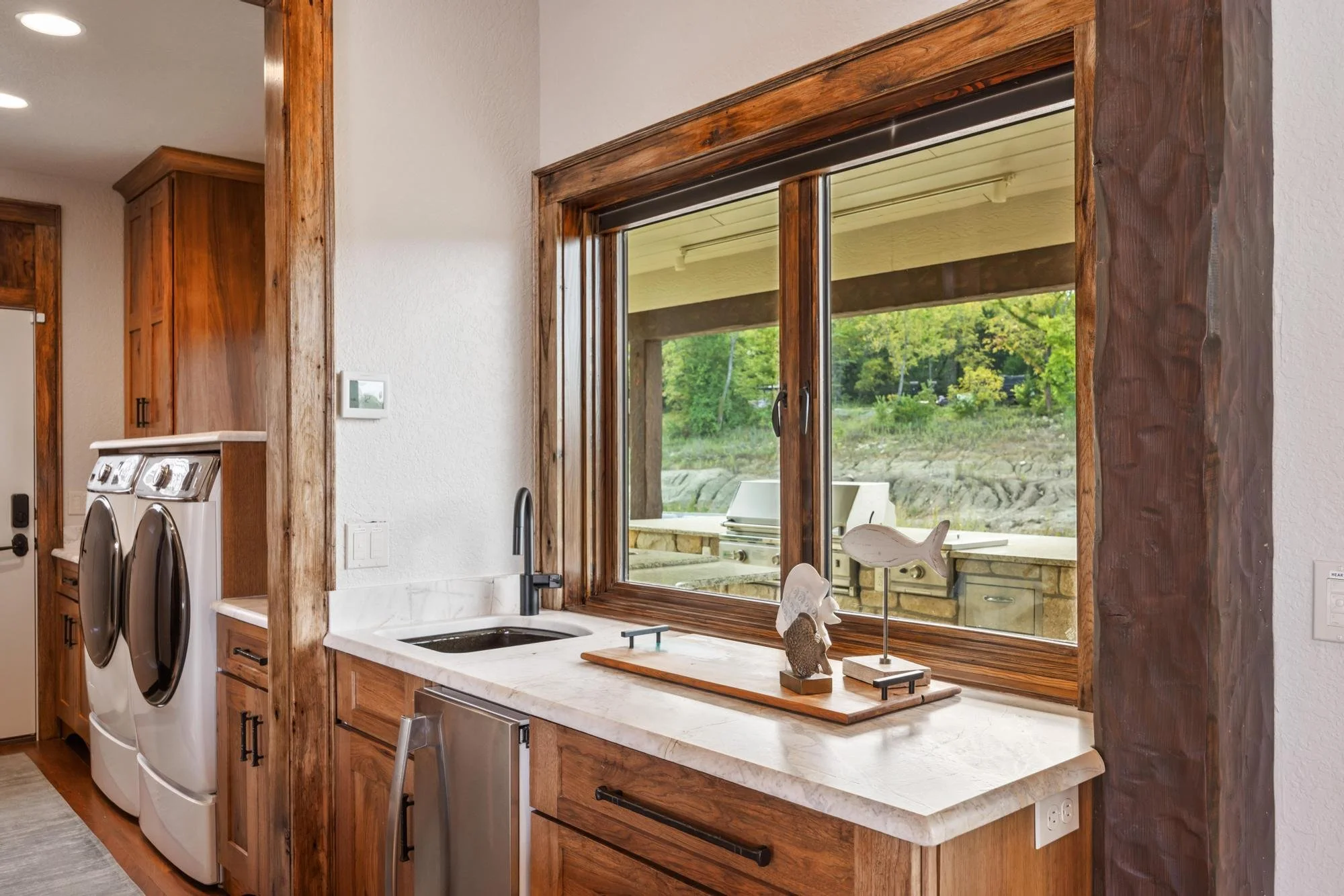 A kitchen with wooden cabinets, a window above the sink, and laundry machines nearby. The outside view through the window shows greenery and rocky terrain.