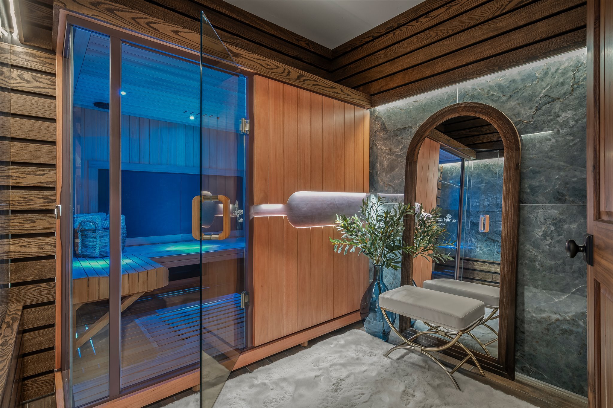 Interior of a modern room featuring a glass-enclosed sauna with blue lighting, wooden walls, and a marble accent wall with an arched opening. There are two white chairs, a small table with a plant, and a soft white rug.