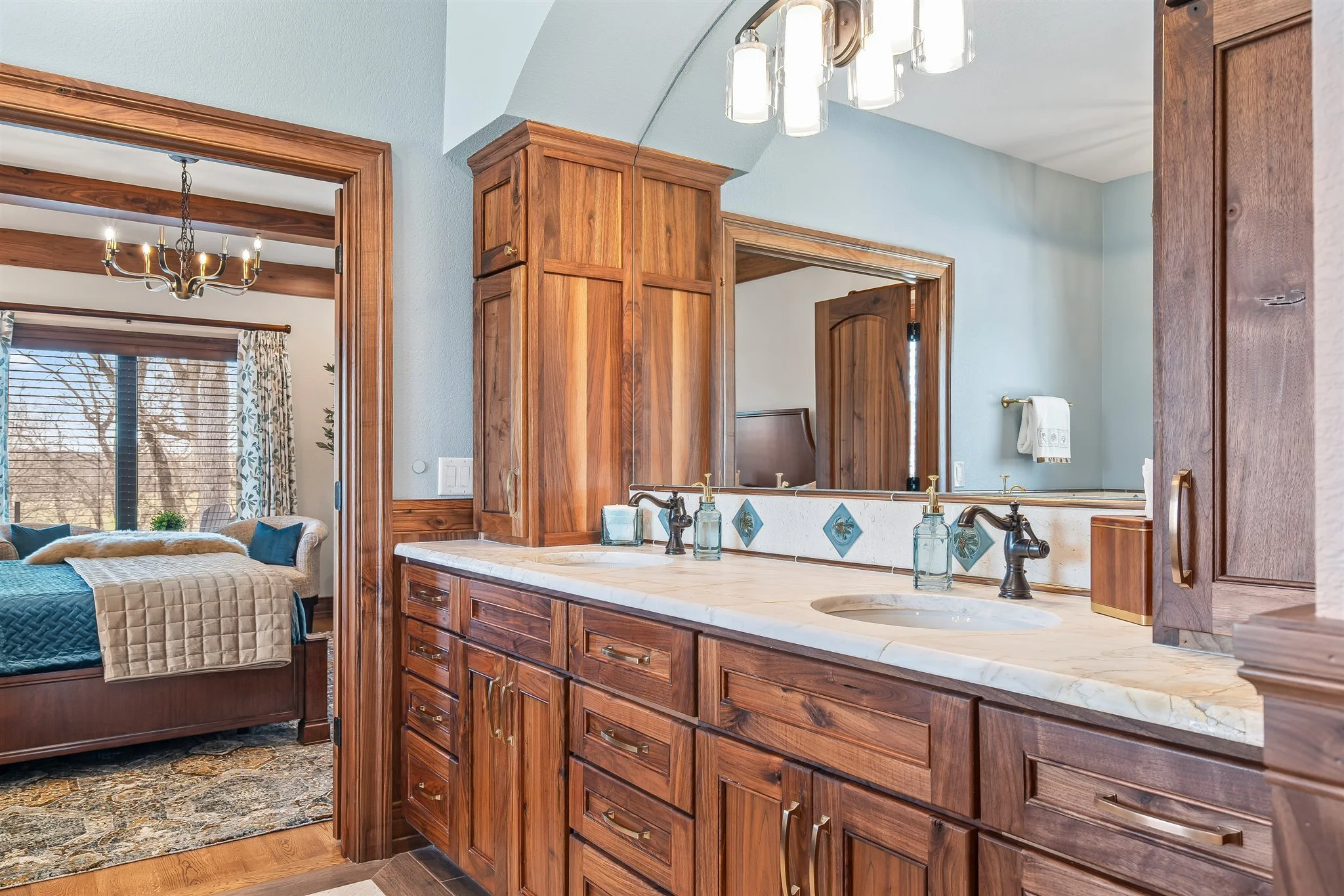 View of a bathroom vanity with wooden cabinets, a marble countertop, and a large mirror, with a bedroom visible in the background featuring a bed, window with curtains, and chandelier.