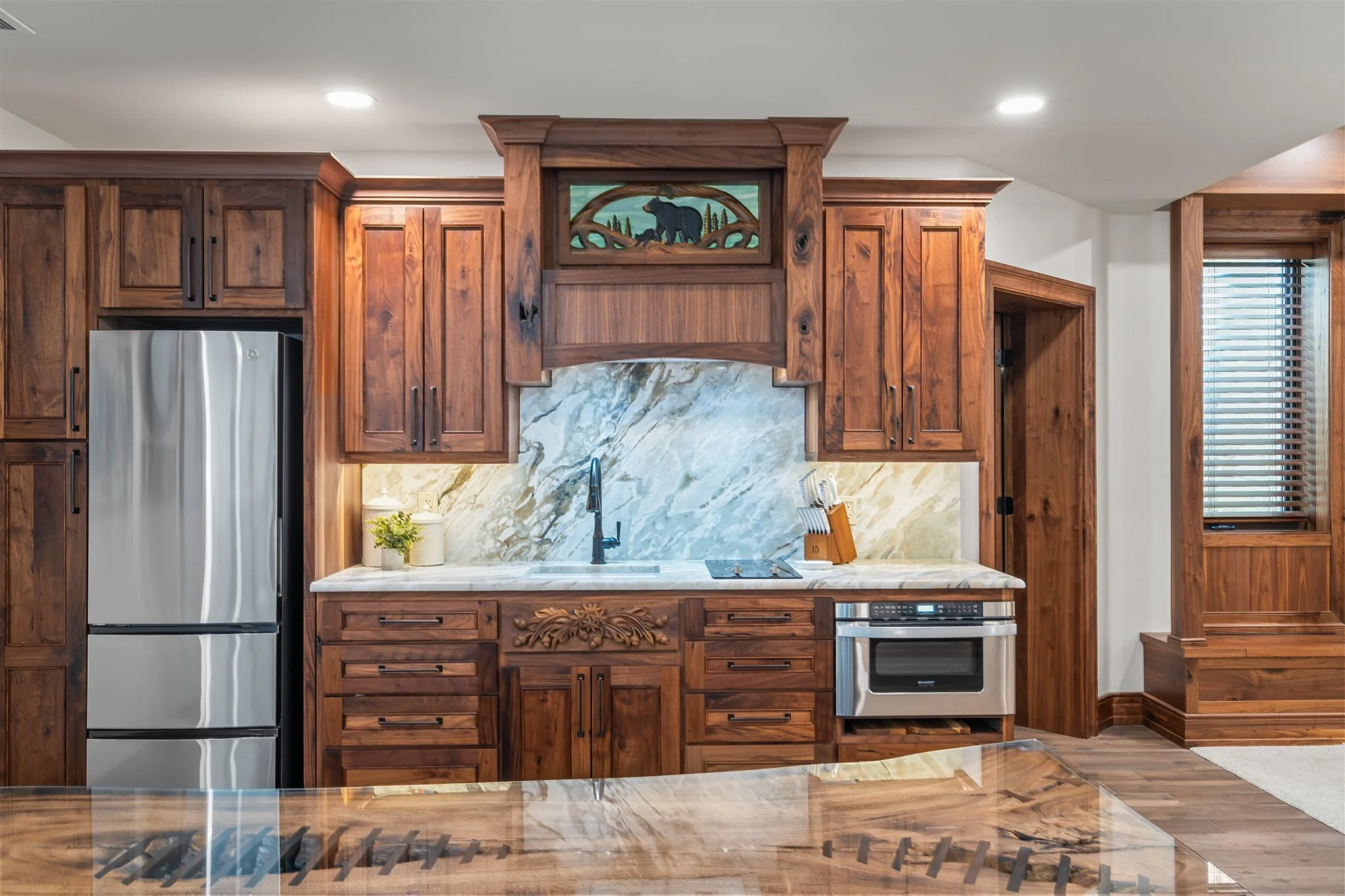 Kitchen with wooden cabinets, marble backsplash, stainless steel refrigerator, and a built-in oven, featuring a decorative bear carving in the upper cabinet area.