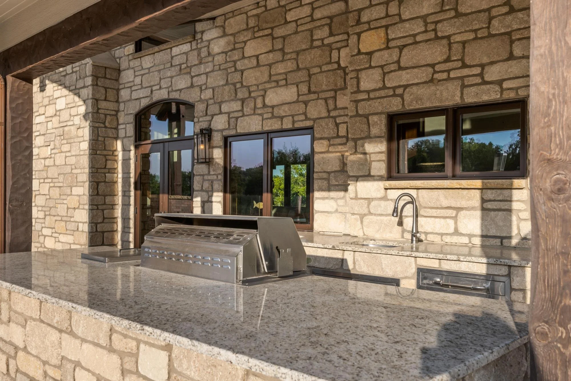 Outdoor kitchen with stone countertop, built-in grill, stainless steel appliances, and windows reflecting trees and sky