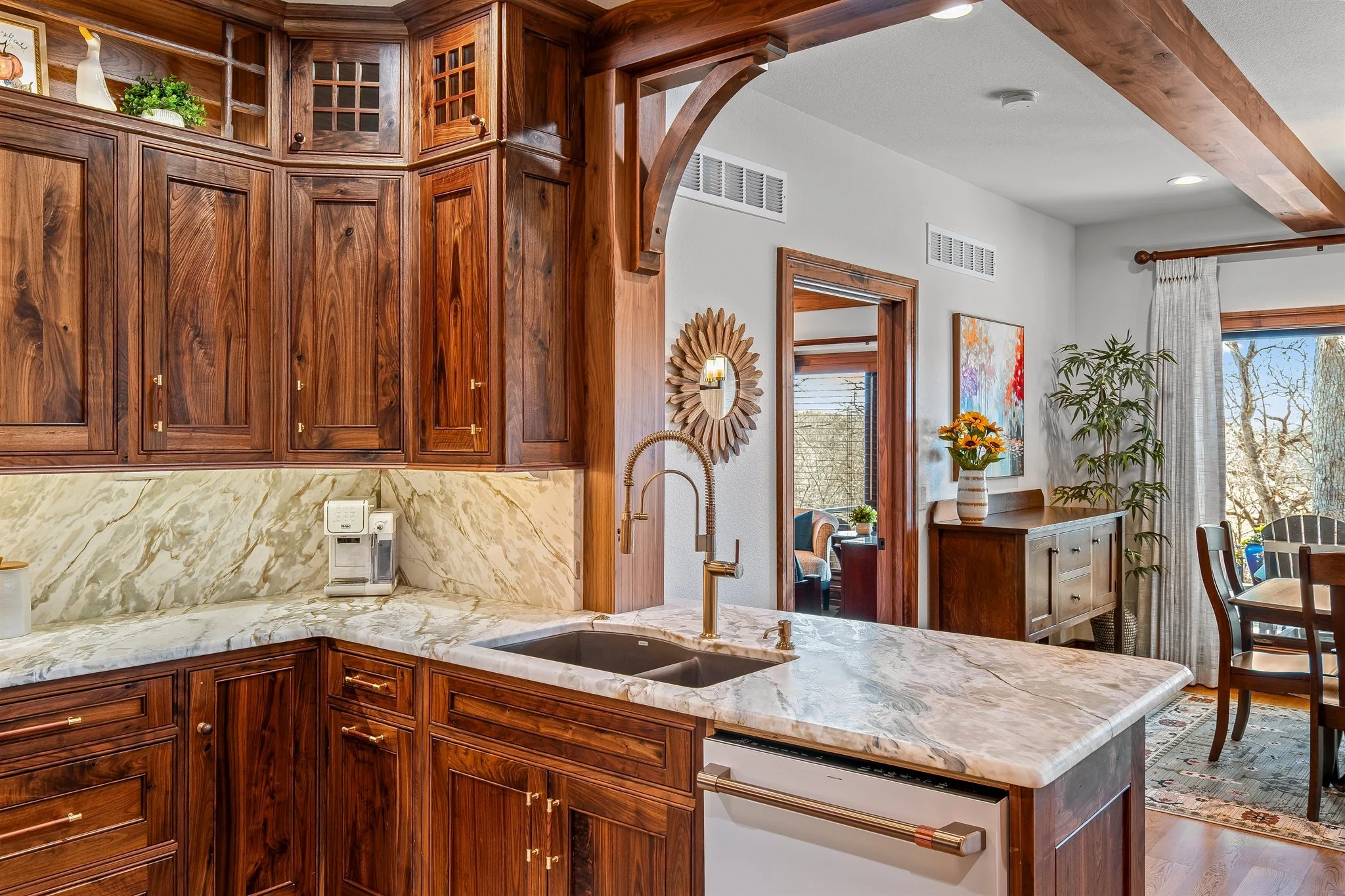 A kitchen with wooden cabinets, a marble countertop, and a double sink with a gold faucet. There is a coffee maker on the counter and decorative items on the shelves. The background shows a dining area with a table, chairs, a vase of yellow flowers, 