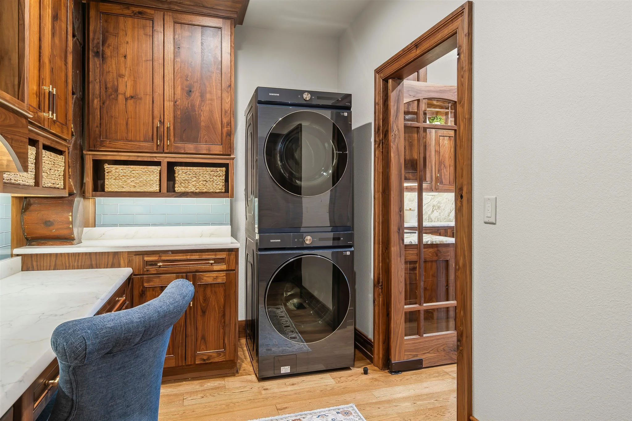 A laundry room with wooden cabinets, a front-loading Samsung washer and dryer stacked vertically, a white countertop, a textured blue chair, and a door with glass panels leading to an adjacent room.