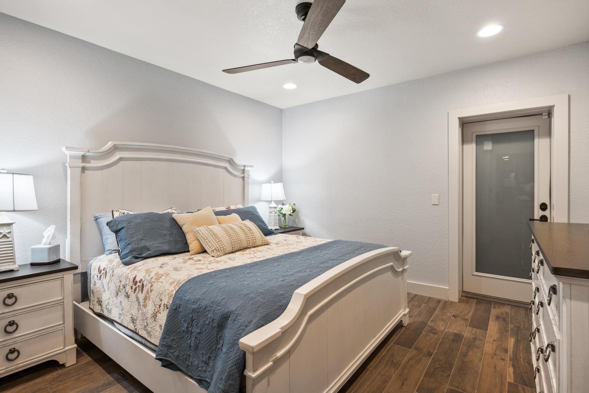A bedroom with a white bed frame, beige and gray bedding, and pillows. There are two nightstands with lamps, a dresser, a ceiling fan, and a door with a frosted glass window. The room has light-colored walls and wooden floors.