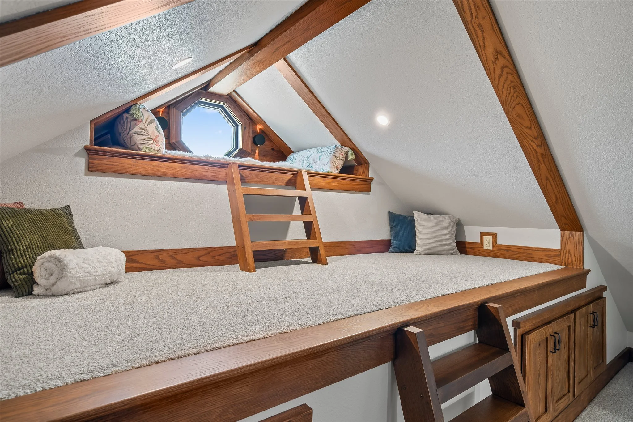 Loft bedroom with a sloped ceiling, wooden beams, a small window, and pillows on a carpeted platform bed.