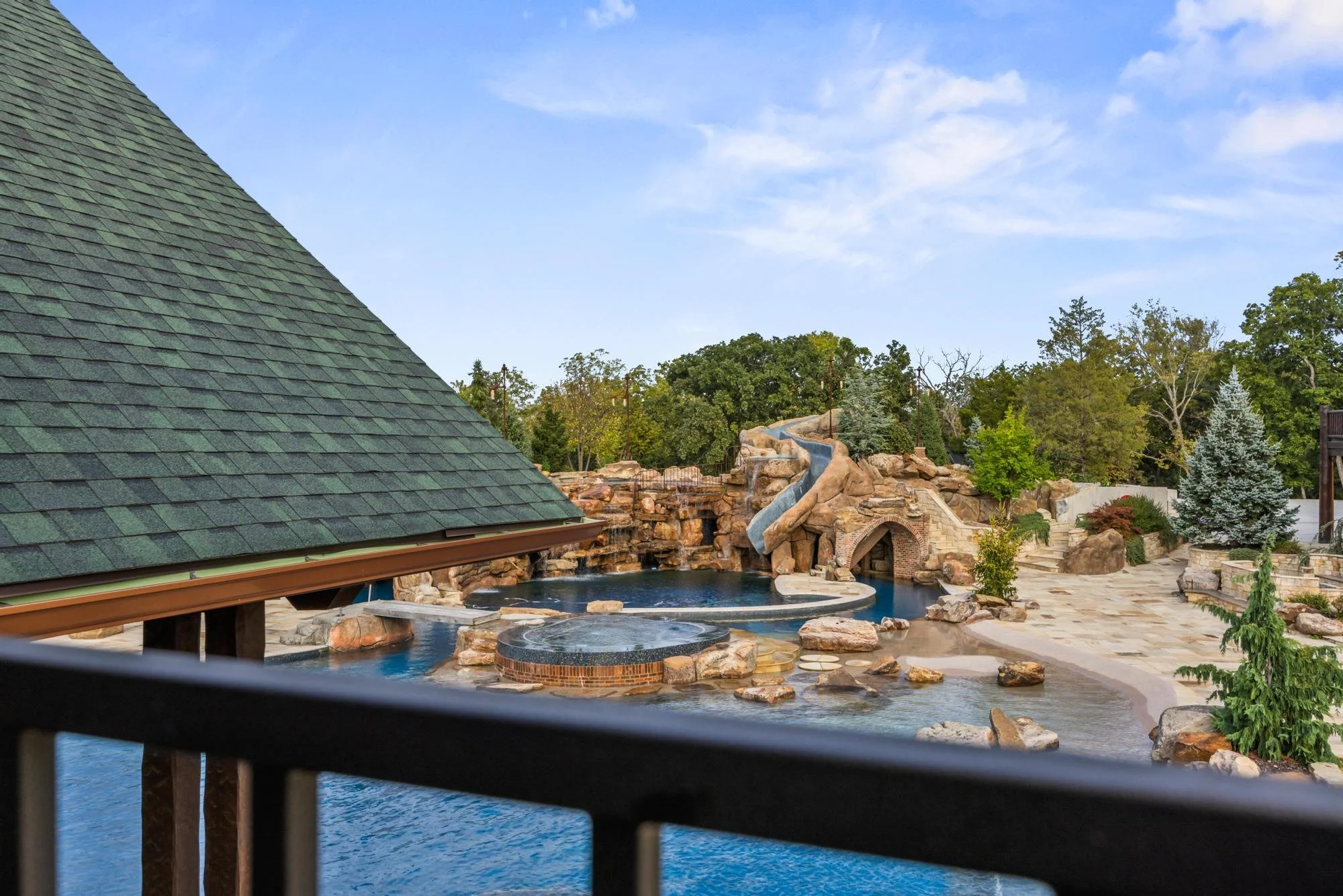View of a backyard with a swimming pool, water slide, rocks, trees, and a house with a green shingle roof, seen from behind a black railing.